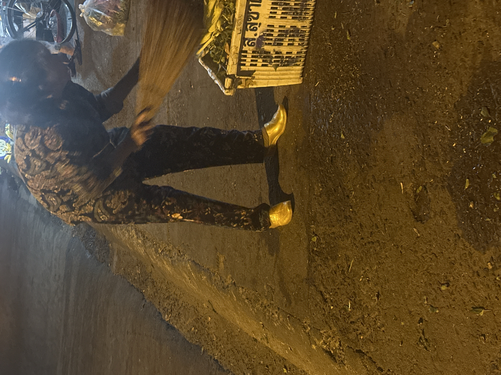 A person with a broom sweeps a street in Bac Nha Trang Ward, Vietnam. The individual wears dark pants, a patterned top, and gold-colored shoes. The street is wet and littered with debris. A bicycle is parked nearby. A plastic basket with text in a local language sits to the sweeper’s side. The lighting suggests it is nighttime.