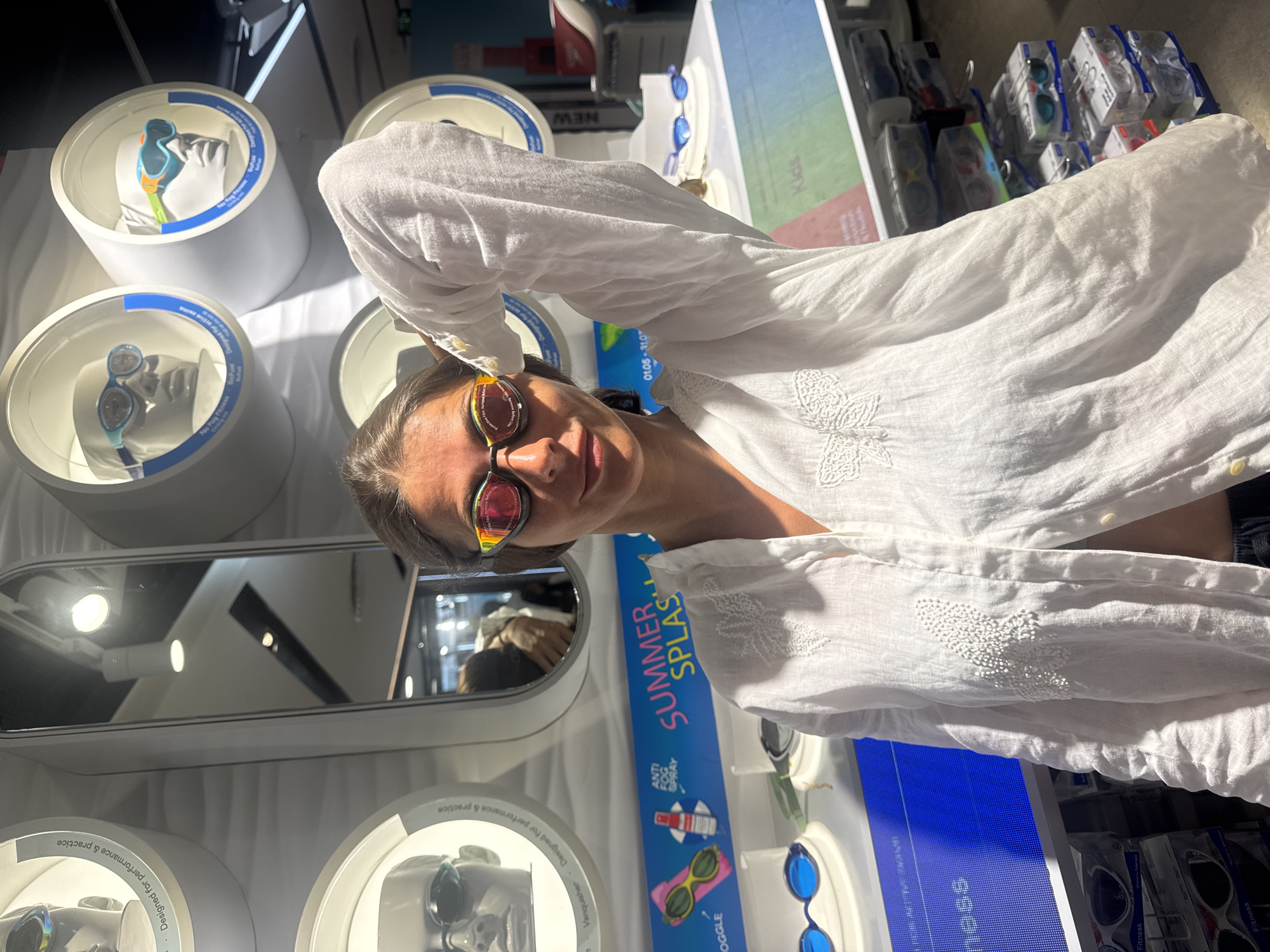 A young woman with dark hair is standing in a store and posing for a photo. She is wearing a white shirt and swim goggles. She is holding her right hand up to her head and looking at the camera. The store appears to be selling swim gear, with displays of goggles and other accessories in the background. One display features mannequins wearing different styles of goggles. There is also a sign that says "SUMMER SPLASH". The woman is positioned in the foreground of the image, while the store's merchandise and displays fill the background.