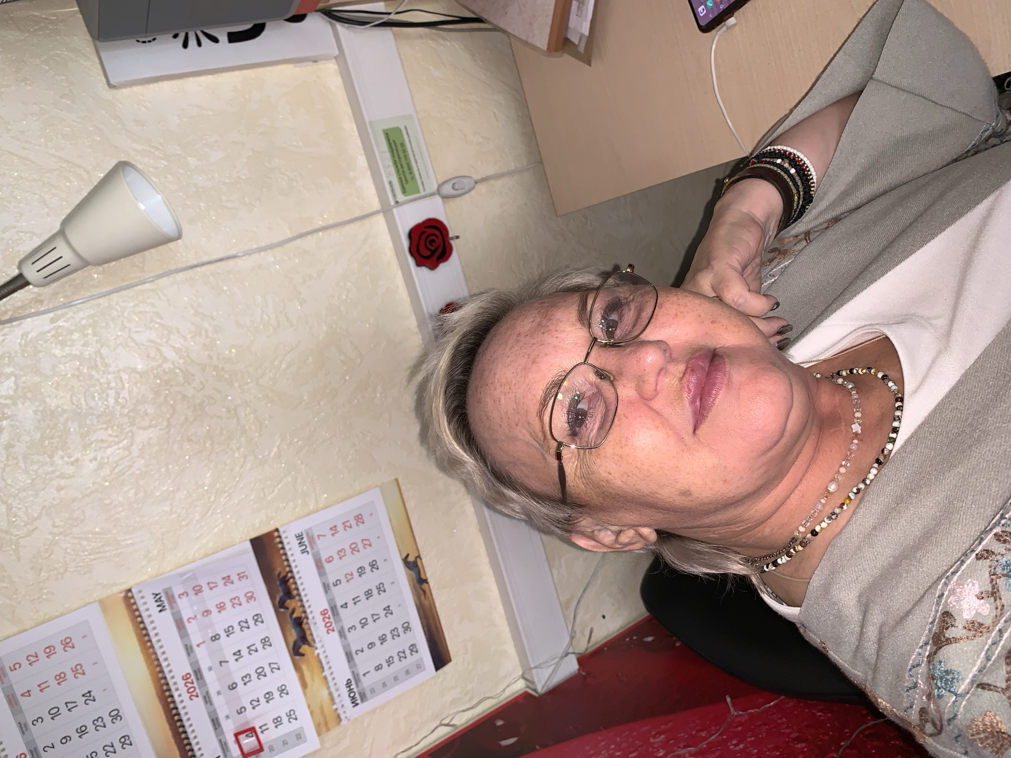 Here's a matter-of-fact description of the image:
**Scene:** An indoor space, possibly a home office.
**People:** A mature woman with blonde hair and glasses is visible. She's wearing a light-colored top under a patterned jacket and several bracelets on her wrist. She is also wearing at least two beaded necklaces.
**Objects:**
* A desk lamp.
* A calendar showing May and June 2026, written in Russian.
* A red rose ornament.
* A smartphone connected to a charging cable on a desk.
**Actions/Interactions:** The woman appears to be resting her head on her hand, looking at the camera.
**Environment:** The room has light colored walls. The visible items suggest an organized workspace.
**Location:** The presence of the Russian calendar suggests this is likely in Russia. Given the script used on the calendars ("Июнь"), we can assume it may be in the city of Chaikovsky, Russia.
