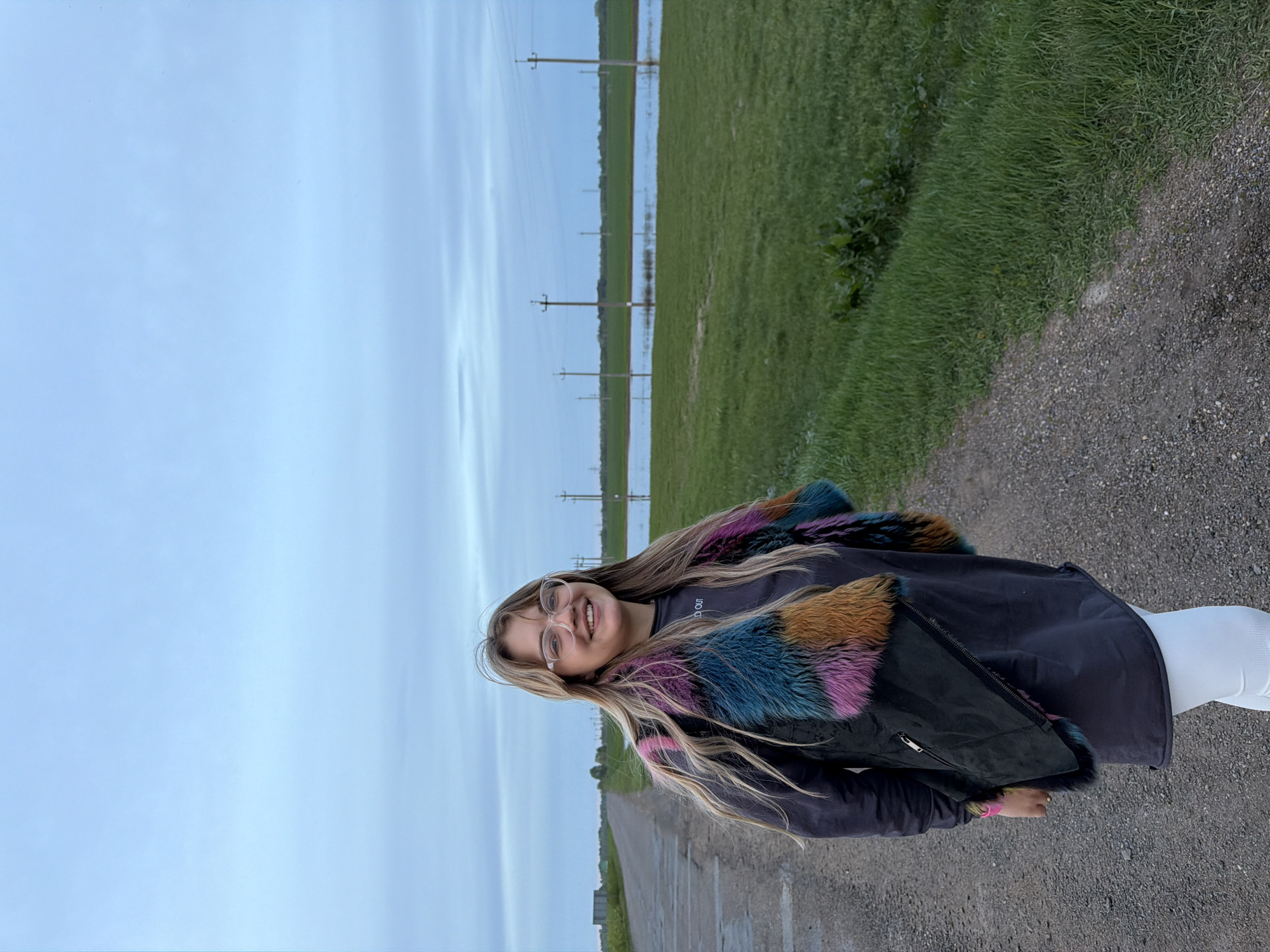 A medium shot depicts a young woman standing on a gravel path in Azyakul', Russia. She is wearing glasses, a dark jacket with colorful faux fur trim, and white leggings. The path stretches into the distance, flanked by a grassy field on one side and a paved road on the other. In the background, utility poles are visible across a flooded area, suggesting recent heavy rainfall. The sky is overcast. The woman is smiling at the camera.
