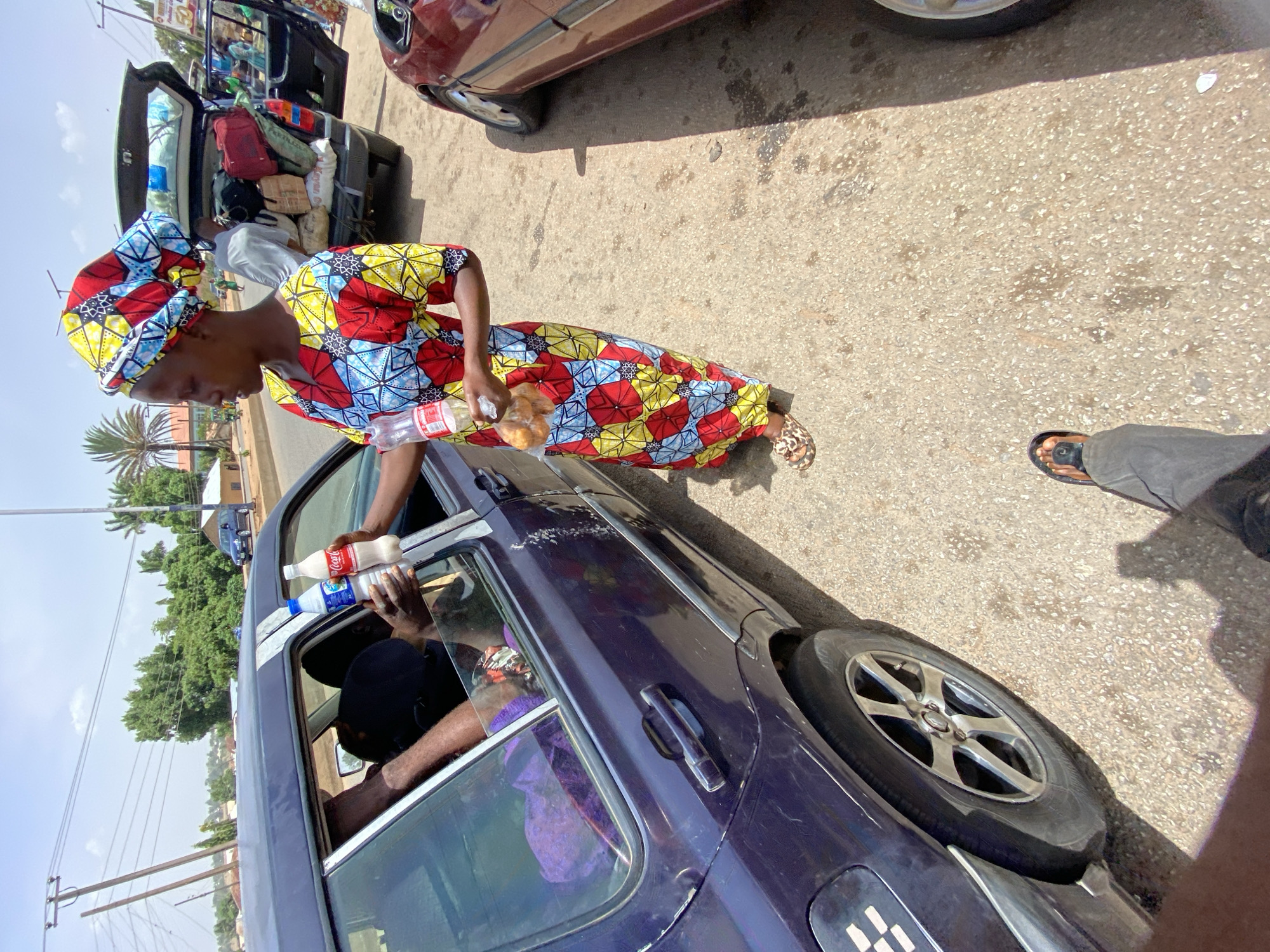 The image, taken in Bukuru, Nigeria, captures a bustling roadside scene under a bright, sunny sky. In the foreground, a woman wearing a vibrant, colorful patterned dress and matching headwrap is engaged in an interaction with a dark blue car. She holds a plastic bottle with a red label, on which "Coca-Cola" is partially visible, and a clear plastic bag containing what appears to be baked goods or bread. Her left hand extends toward the car's open window, where two hands from inside the vehicle are visible, holding two other plastic bottles—one white/milky and one clear with a blue cap—suggesting a transaction or exchange of items.
Behind the woman, another dark-colored car, possibly a sedan or SUV, has its trunk wide open and heavily laden with a variety of luggage, including bags, sacks, and boxes, indicating preparations for or the end of a journey. A person is partially visible near the trunk. To the right of this car, part of a red/brown vehicle is also seen. The ground is paved, and the general setting includes utility poles with power lines, lush green trees (including a prominent palm tree), and faint structures in the distance, typical of an urban or semi-urban environment. The time of day is clearly daytime, with strong sunlight and visible shadows. The scene depicts a common sight of roadside vendors and travelers in the region.