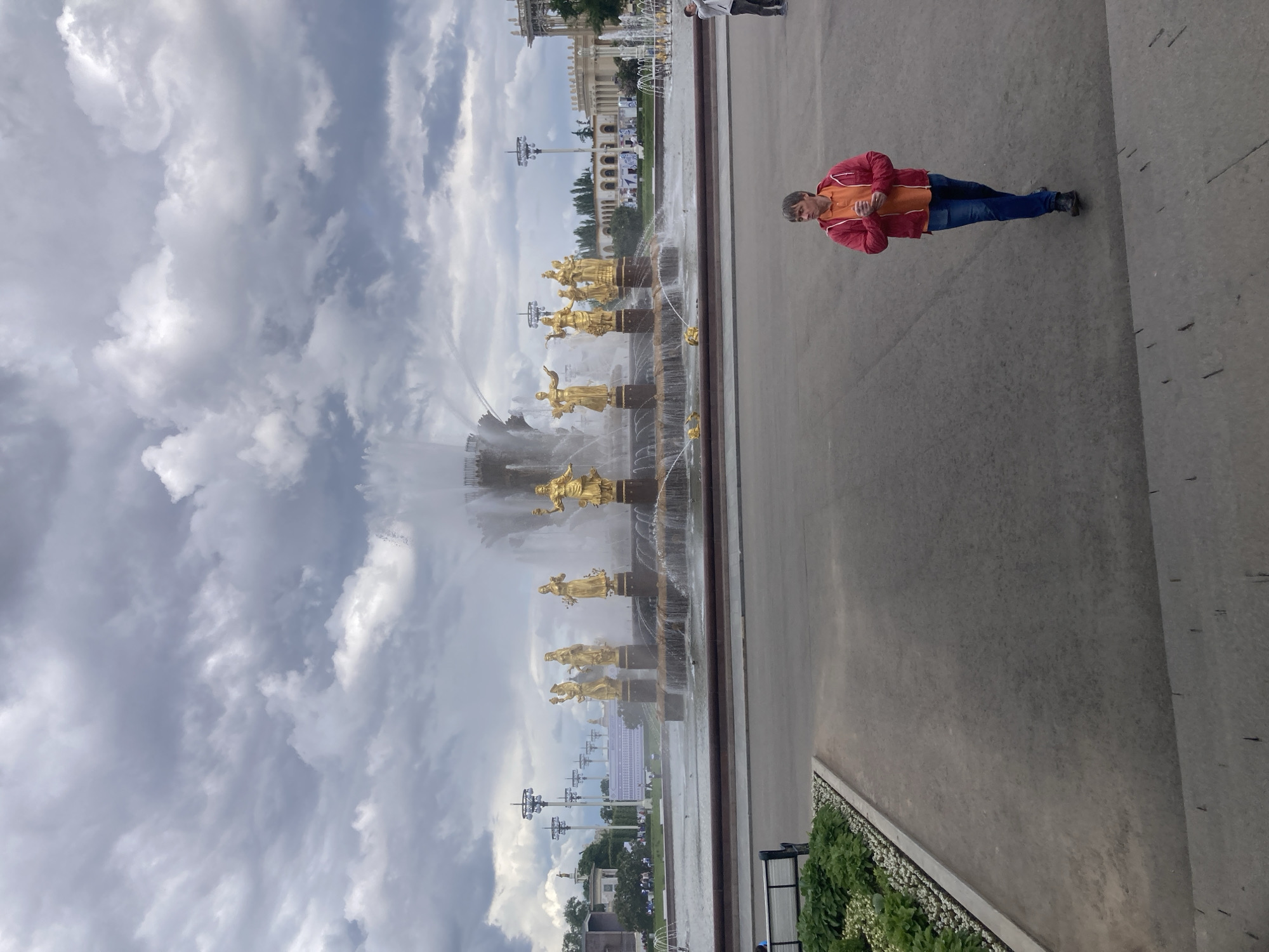 A medium shot captures a person standing on a concrete surface next to a long fountain featuring a series of golden statues. The individual, dressed in an orange shirt and a red jacket, is also wearing blue jeans and appears to be looking at their phone. The fountain is the "Friendship of Nations" fountain, situated in the VDNKh park in Moscow, Russia. The statues represent the former Soviet republics. The sky above is filled with dramatic, cloudy formations. Some buildings and streetlights are also visible in the background.
