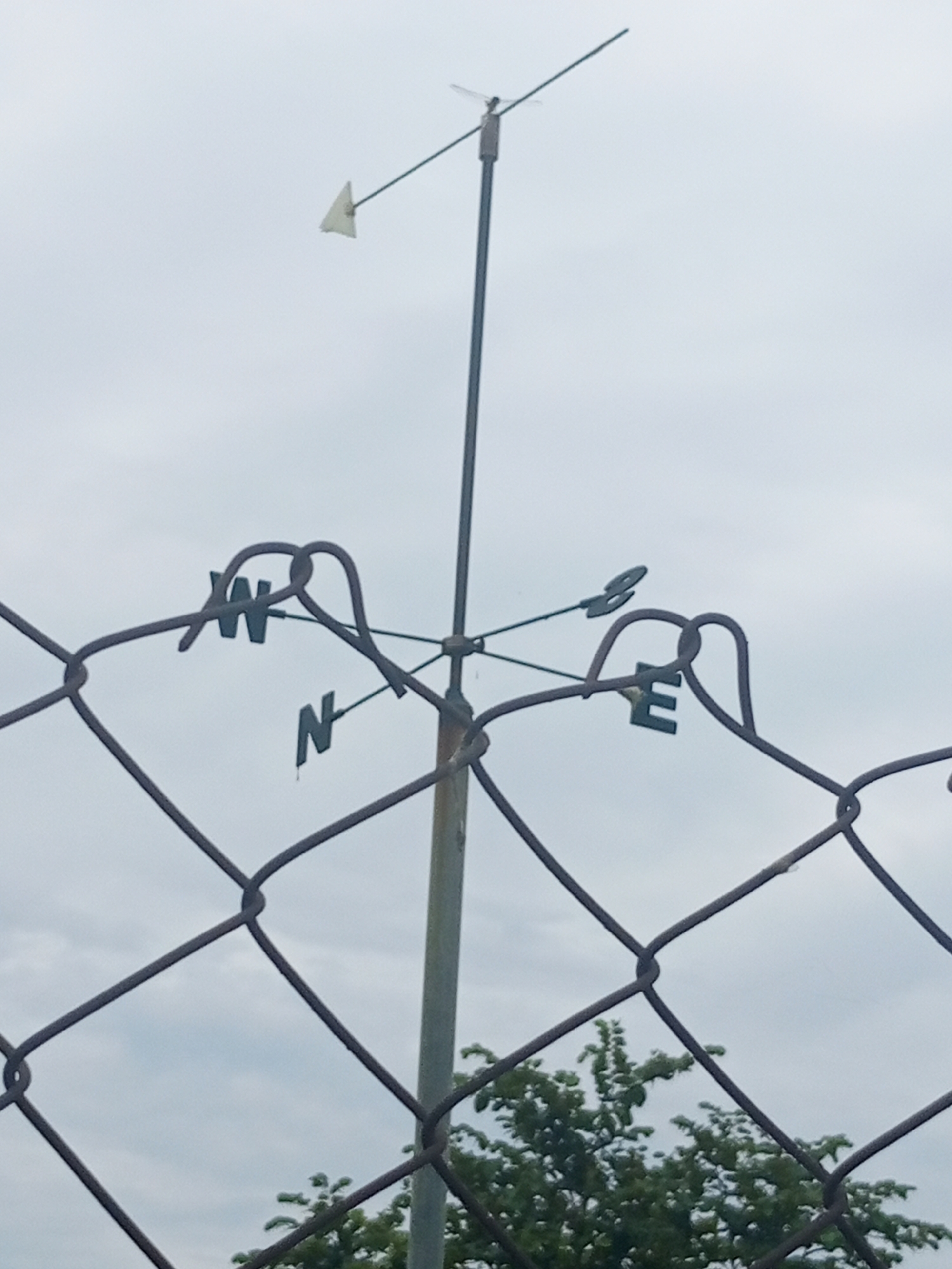 A close-up, outdoor, eye-level shot shows a weather vane attached to a pole, positioned behind a chain-link fence in Jalingo, Nigeria. The weather vane has directional letters 'W', 'N', and 'E' attached to it, with the 'N' pointer facing downwards, indicating that the wind is blowing from the north. A small, triangular flag is attached to the weather vane and points to the west. The sky is overcast and gray. In the background, green foliage from trees is visible. The chain-link fence occupies the foreground of the image, partially obscuring the view of the weather vane and the background.