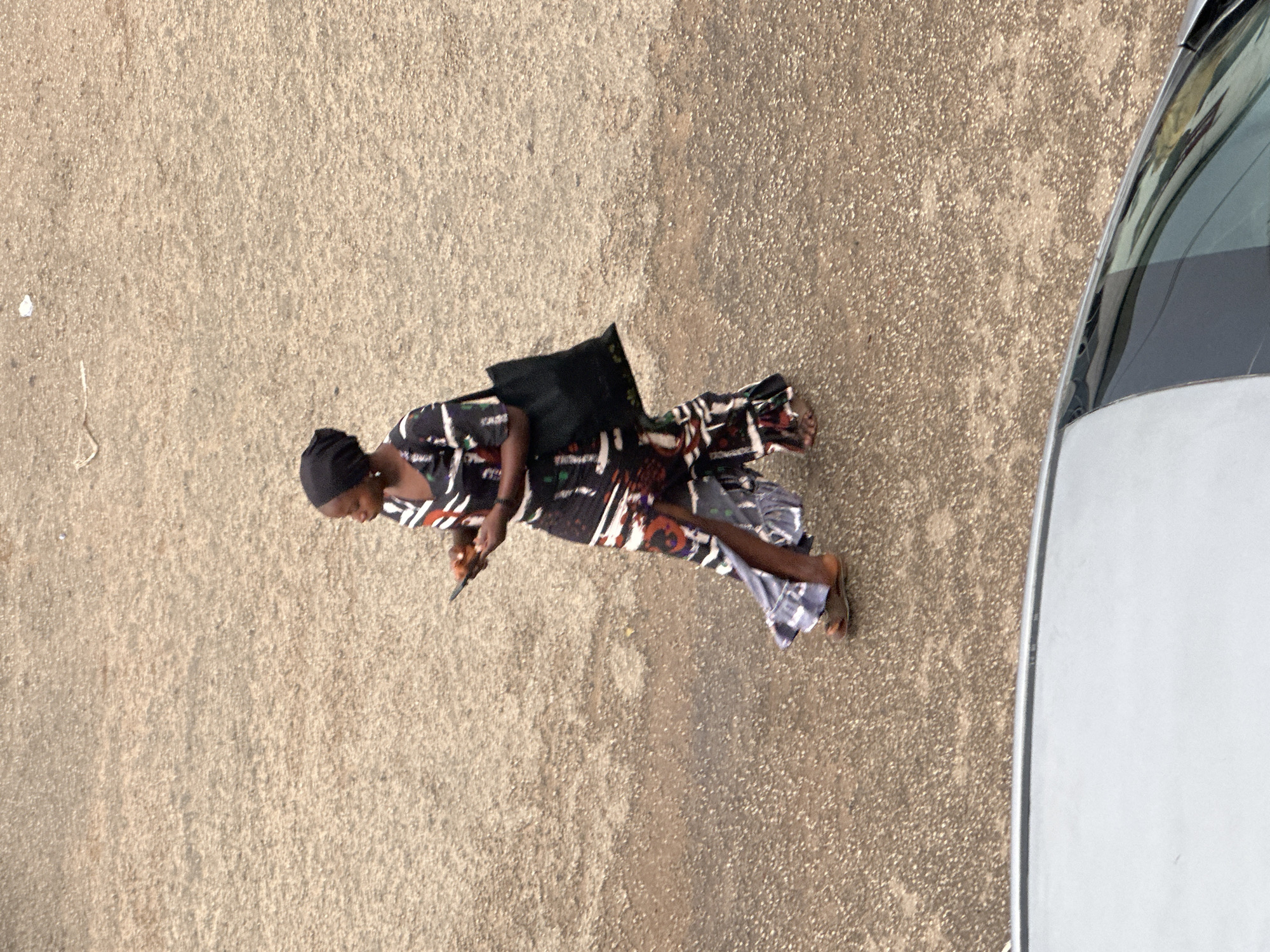 A lone Black woman is walking on a dirt road in Bukuru, Nigeria. She is wearing a dark headscarf, a patterned dress with a slit up the side, and sandals. She is carrying a black tote bag over her right shoulder and holding a mobile phone in her left hand. The road surface is textured and speckled with small stones and dirt. A portion of a light-colored car is visible in the foreground, its windshield reflecting the surrounding environment.