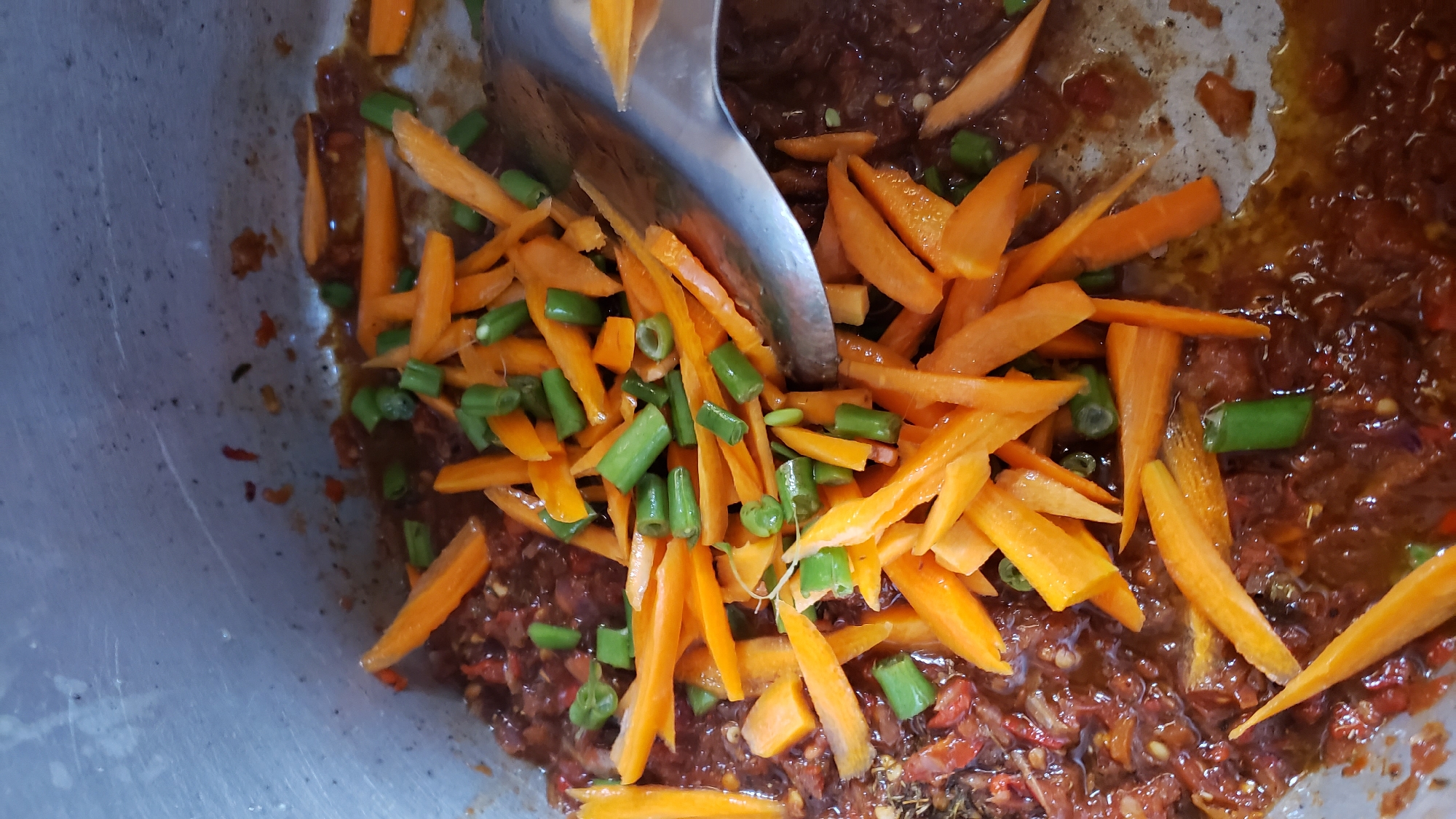 A close-up, overhead shot shows chopped carrots and green beans being added to a pot of stew. The stew, a deep reddish-brown color, is being stirred with a large metal spoon. The pot itself appears to be made of a dull metal, possibly aluminum, and the interior is somewhat stained. The scene is consistent with food preparation taking place in a home kitchen in Narayi, Nigeria, a residential area. The image focuses entirely on the food and the cooking process, with no discernible background elements that would provide further environmental context.