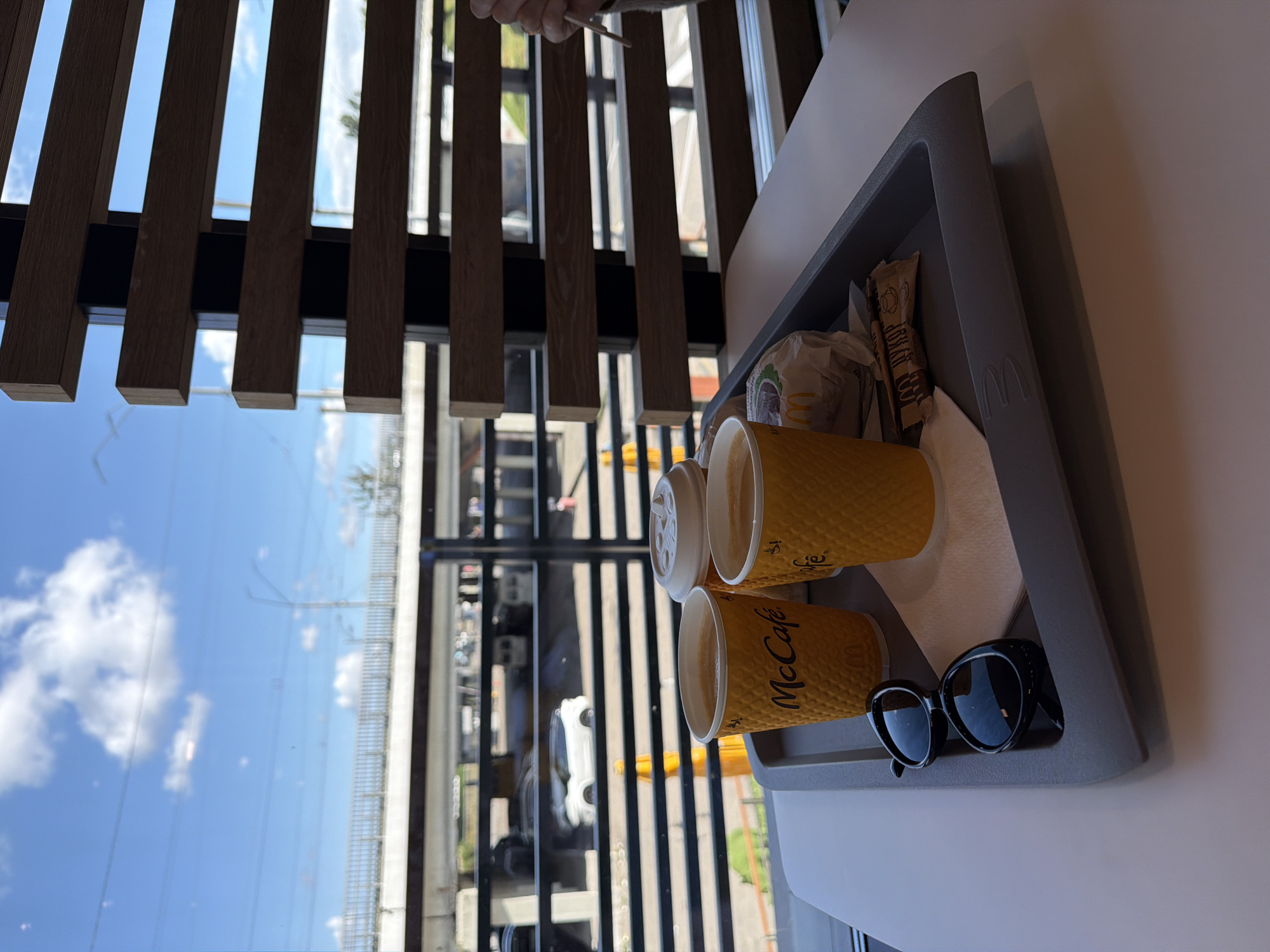 An outdoor dining scene is presented, viewed through a window with wooden slatted blinds. A gray tray sits on a white table, holding two McCafe coffee cups, a wrapped burger, sunglasses, and a packet of sugar. Outside the window, a highway overpass is visible with cars driving on it. The sky is blue with scattered white clouds. The scene is likely in Kyiv, Ukraine.