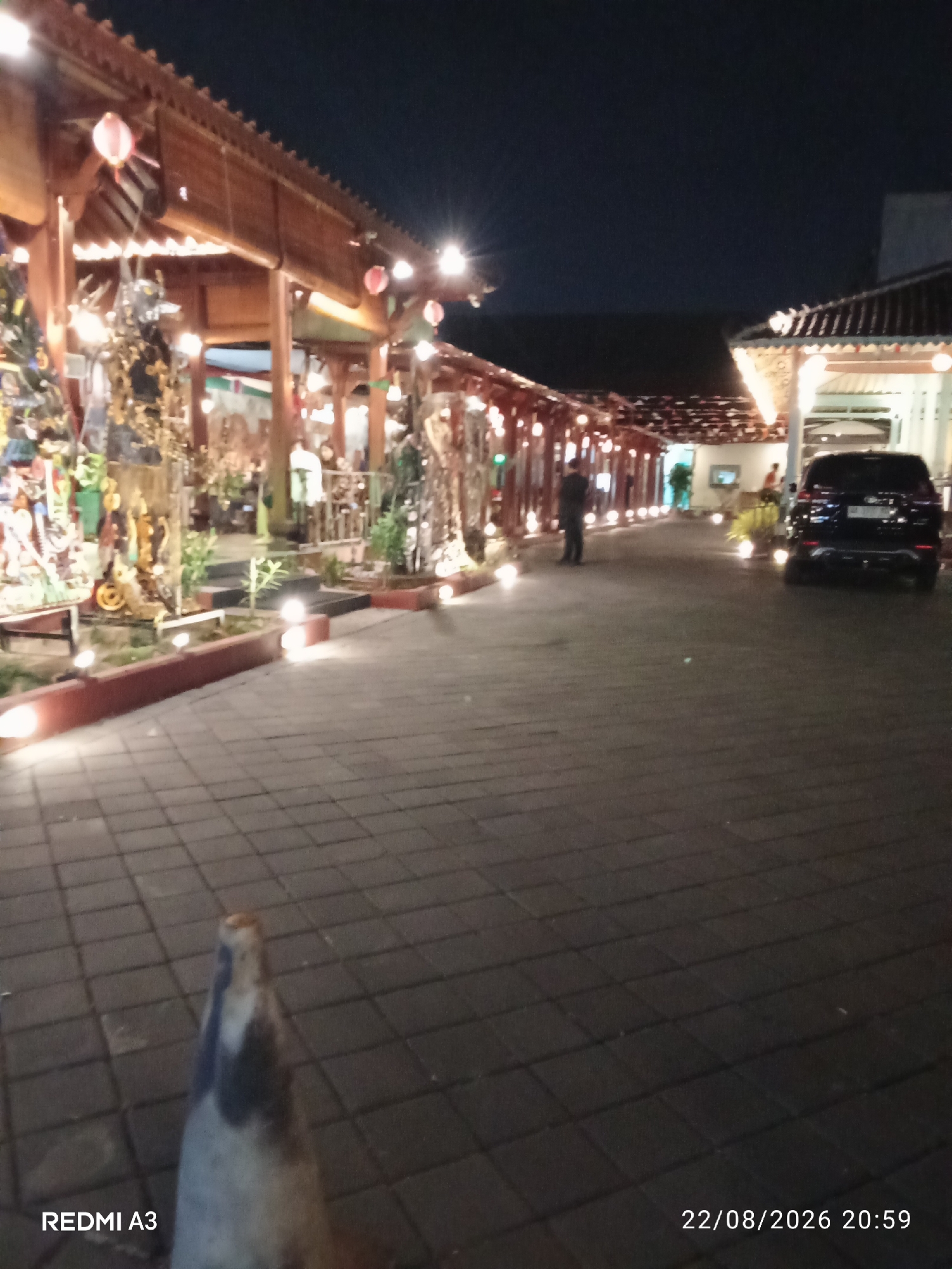The image captures a nighttime scene in Surakarta, Indonesia, likely at an outdoor venue or restaurant. The foreground shows a tiled walkway. On the left, there is a covered walkway with wooden architecture, adorned with decorative elements and illuminated by lanterns and string lights. Various sculptures and decorative items are visible within this area. Further into the scene, a dark-colored SUV is parked on the right side. Several people are present, appearing to be walking or standing. The overall ambiance is one of a well-lit, possibly festive, evening setting. The bottom left corner displays "REDMI A3," and the bottom right corner shows the date and time "22/08/2026 20:59."