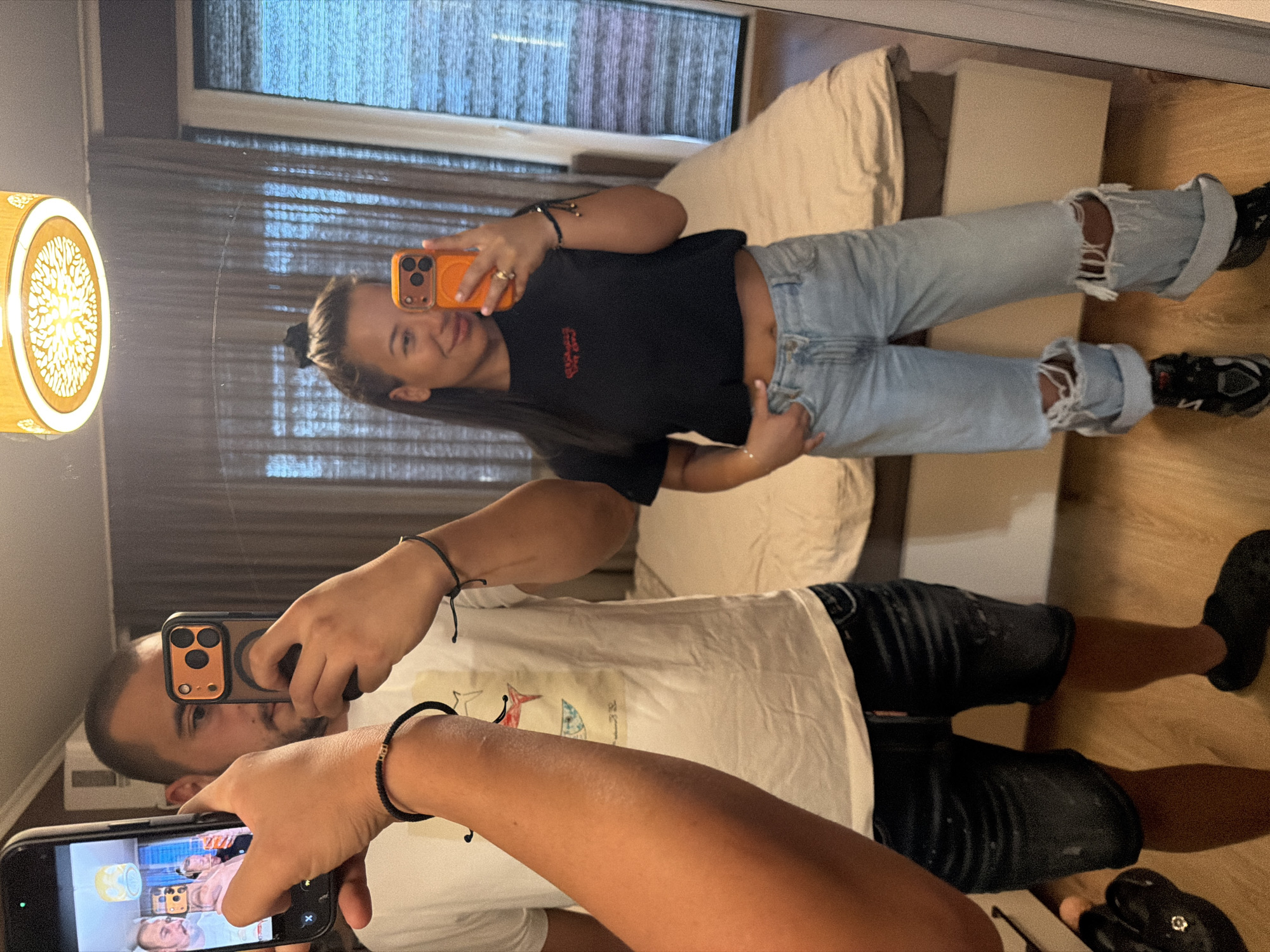 In a domestic setting, likely a bedroom in Burgas, Bulgaria, a man and a woman are taking a mirror selfie. The man, positioned on the left, is holding a smartphone in his left hand, with his right hand holding another smartphone up to the mirror. He is wearing a white t-shirt with a graphic print and dark, distressed denim shorts. The woman, on the right, is also holding a smartphone, a bright orange one, and is looking into the mirror with a slight smile. She is wearing a black crop top with red lettering and light-wash, ripped denim jeans that are rolled up at the cuffs. The background reveals a bed with beige bedding, draped curtains, and a textured ceiling light fixture. The floor is made of wood. The overall scene suggests a casual, personal moment captured within a residential space.