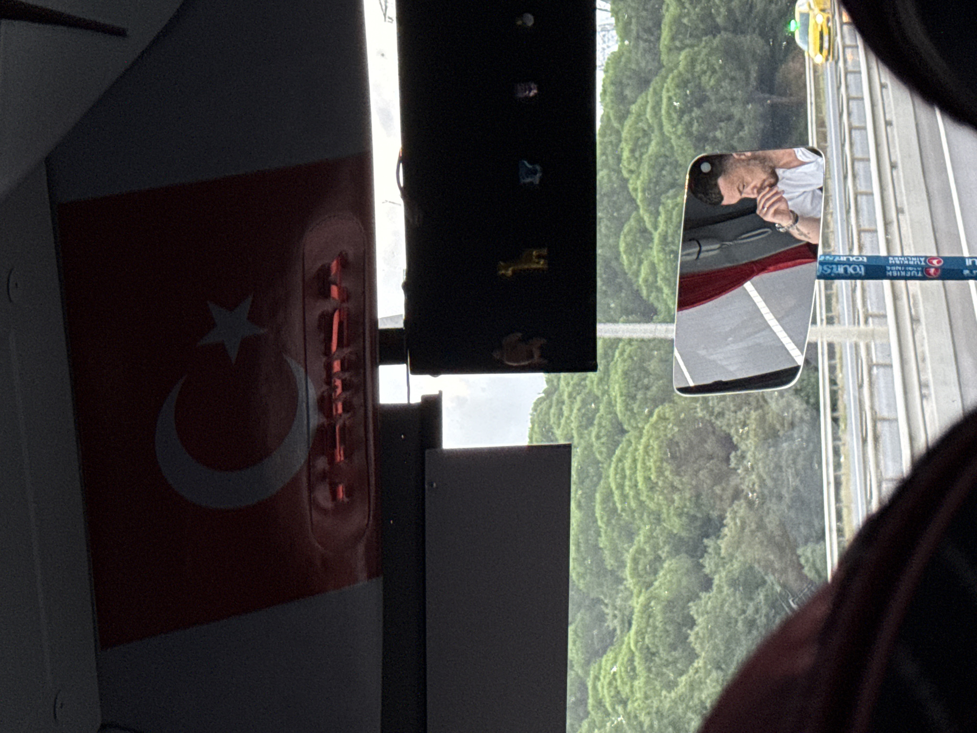 The image appears to be taken from inside a vehicle, possibly a bus, looking out through the windshield and a side mirror. In the upper portion of the frame, a Turkish flag with a crescent moon and star is visible, affixed to the interior ceiling of the vehicle. Below the flag, a dark rectangular panel is mounted.
Further down, a shelf-like structure holds several small decorative figurines, including what appears to be a bear, a giraffe, and other unidentifiable shapes. Beyond this, through the windshield, a dense forest of evergreen trees forms the backdrop.
A side-view mirror reflects the image of a man with short dark hair, wearing a white t-shirt, with his hand near his face. He appears to be contemplating or in deep thought. He has tattoos on his arm. In the background of the mirror's reflection, a red curtain is visible, along with parts of the vehicle's interior.
Outside the vehicle, a highway can be seen with metal guardrails. A bright yellow taxi cab is driving on the highway. Two blue and white signposts with the word "TOURIST" and the Turkish Airlines logo are also visible along the side of the road. The overall lighting suggests it is daytime, possibly overcast. The scene does not explicitly indicate the city of Eyüpsultan, Turkey, but the presence of the Turkish flag and Turkish Airlines branding suggests a location within Turkey.