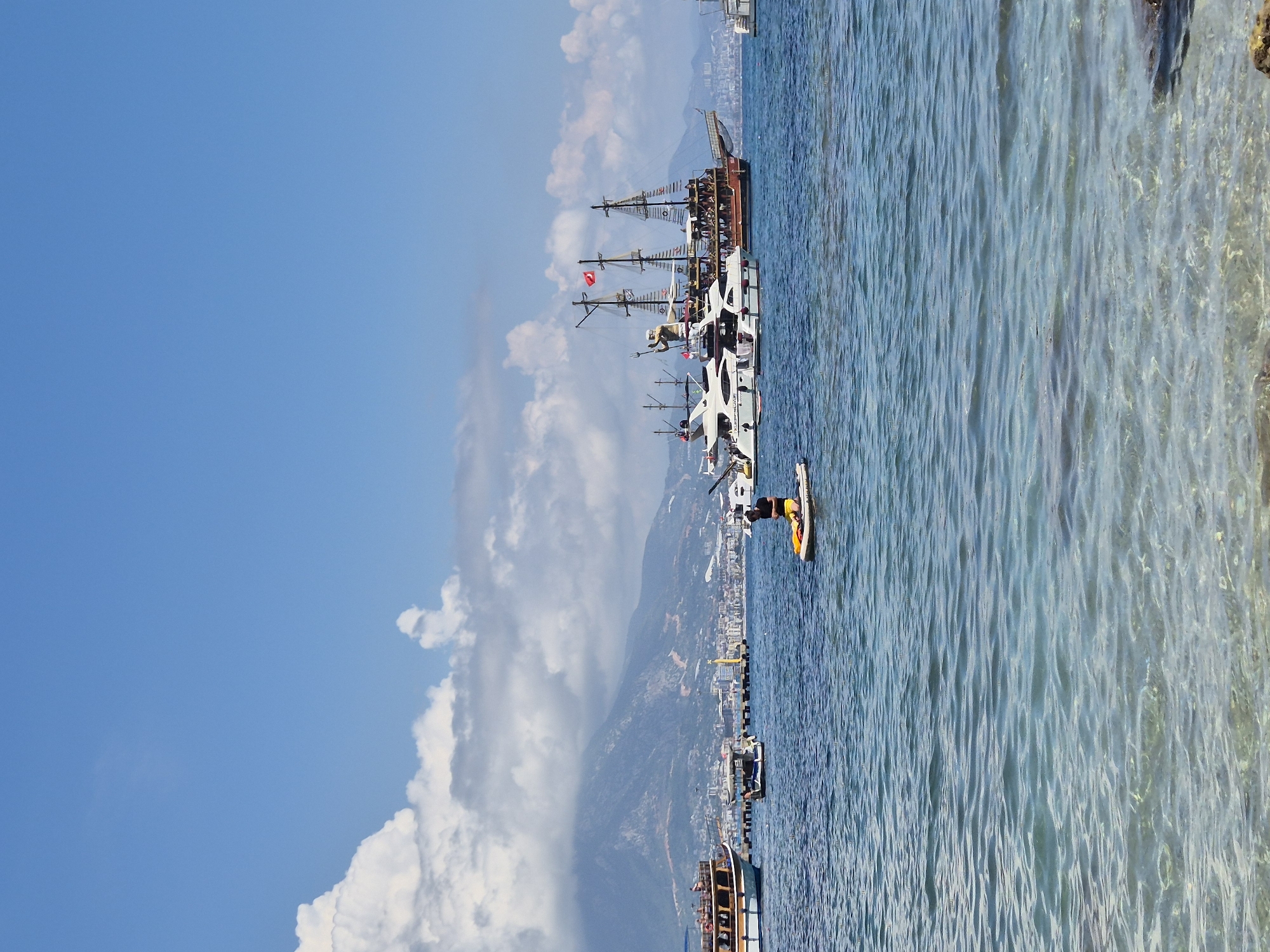 In Alanya, Turkey, a person is paddleboarding on clear blue water. In the background, several boats are anchored, including a large pirate ship and a modern yacht. A town with buildings and mountains can be seen further in the distance, under a partly cloudy sky.