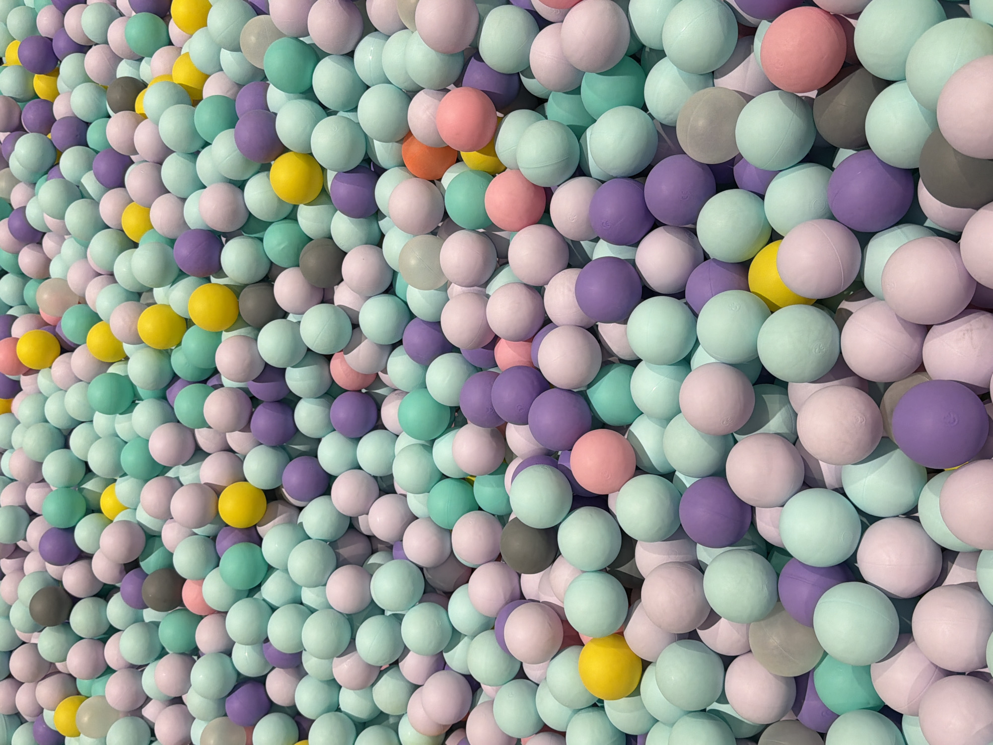 The image shows a dense, wall-like accumulation of pastel-colored balls, primarily in shades of light pink, mint green, lavender, and pale yellow, with some gray and white balls interspersed. The balls are tightly packed, creating a textured surface. They appear to be made of plastic or a similar material and are roughly spherical. The lighting suggests an indoor setting, possibly a ball pit or a decorative display. The overall impression is one of playful, soft colors and a sense of abundance. No people are visible in the image, and no specific activity is depicted beyond the static arrangement of the objects. The time of day, weather, or specific location cues are not discernible from the image itself. There is no visible text in the image.