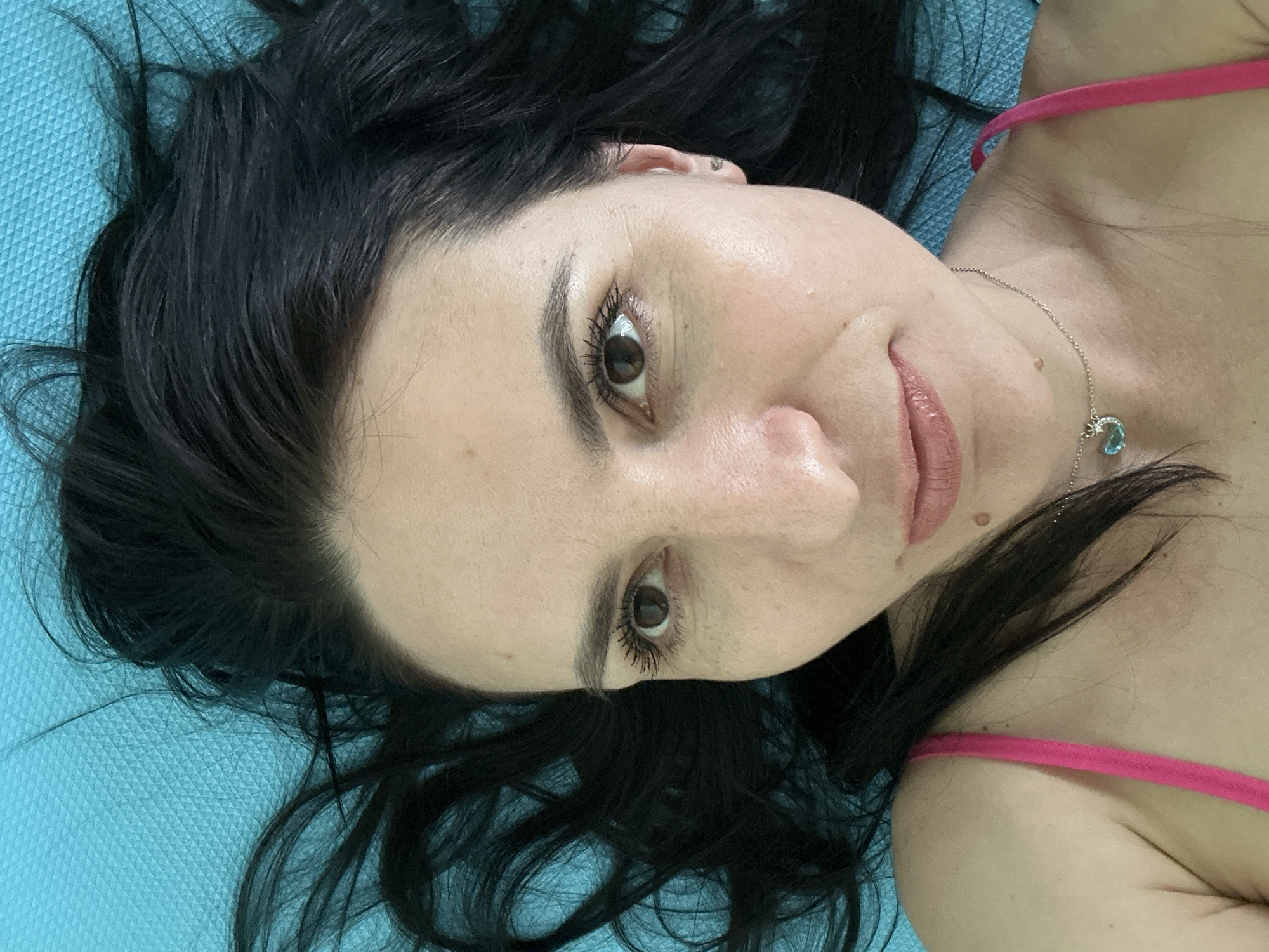 A woman with dark hair is lying on a blue surface. She is wearing a pink top and a silver necklace with a light blue pendant. Her face is visible, and she appears to be looking at the camera. There is no indication of location in a city.