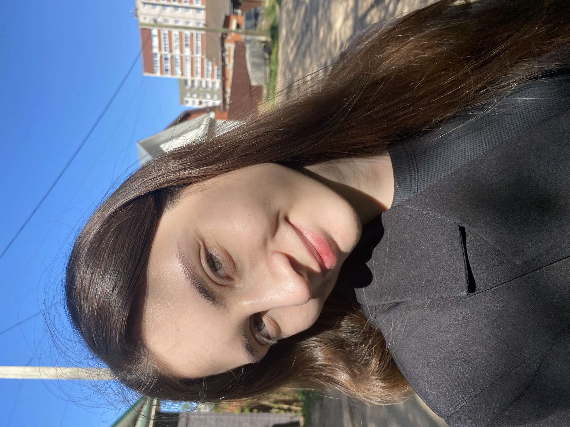 A woman with long, brown hair is shown in a close-up selfie outdoors. She has fair skin, visible eyebrows, and light-colored eyes. She is wearing a black top or jacket. The background shows a street setting with residential buildings under a clear, blue sky. In the background, an apartment building with white and red brick facade suggests the location is in a city in Russia, likely Votkinsk.