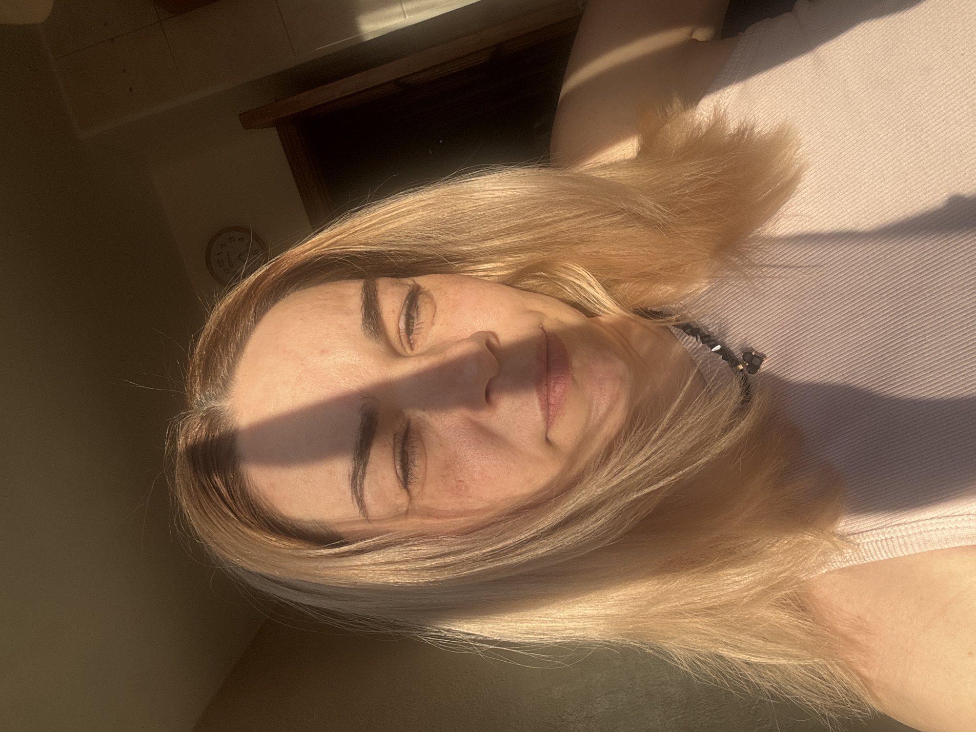 A woman with blonde hair and closed eyes is lying on her back in a room. The room appears to be indoors, judging by the partial view of a clock on the wall and the indoor lighting. She is wearing a light pink top, and a portion of a necklace is visible near her neck. A strong light source casts a shadow across her face. The location is not discernible from the image.