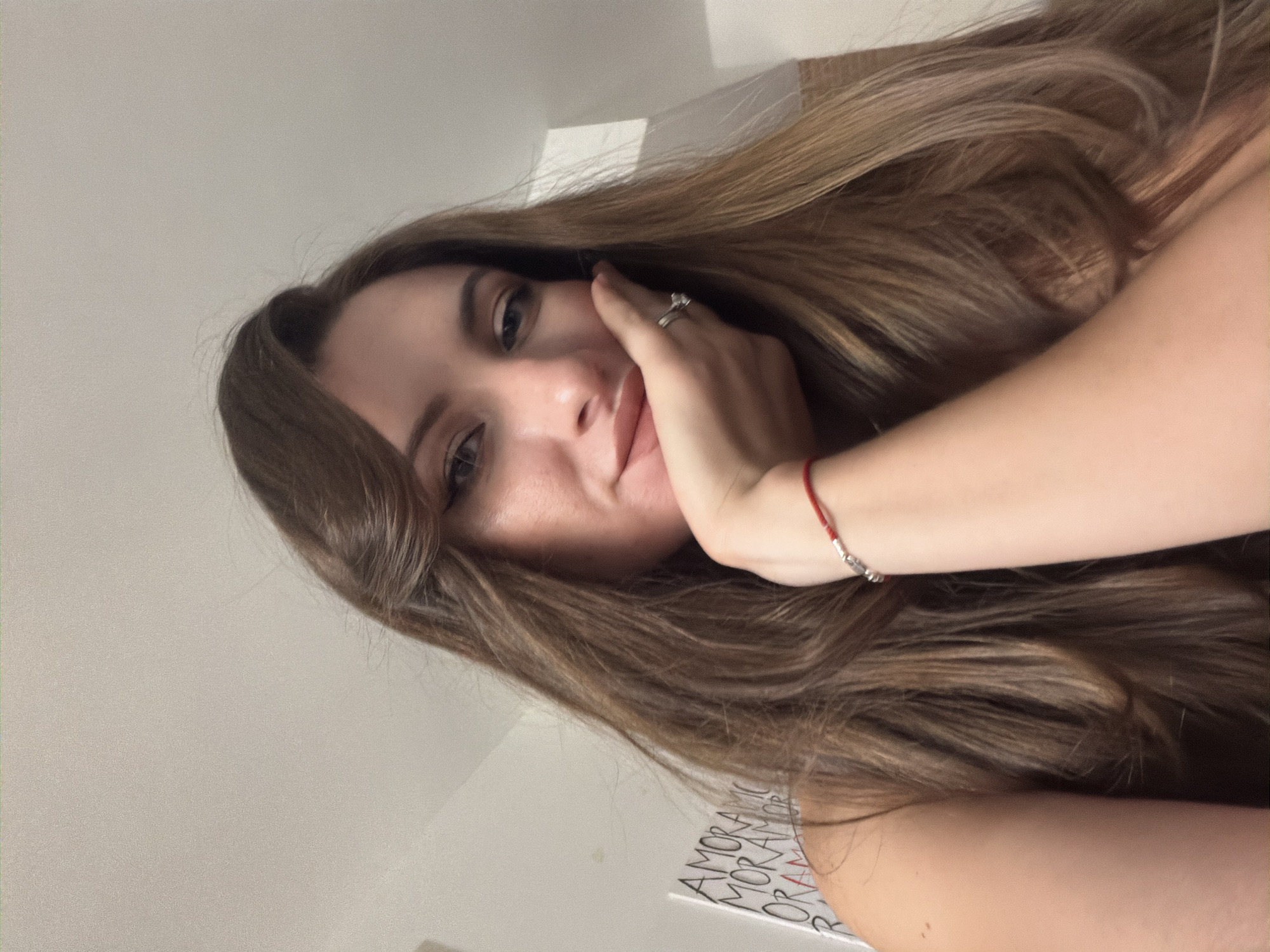 In an indoor setting, a young woman with long, brown hair is captured in a close-up shot, resting her chin on her hand. She is wearing a ring on her right hand and a thin red bracelet on her wrist. Her expression is soft and slightly tilted, with her gaze directed towards the viewer. The background appears to be a wall with some text visible, and the lighting suggests an ambient indoor environment. The overall scene is intimate and personal, focusing on the subject's facial features and pose. No elements in the image suggest a specific location in Sharm El Sheikh, Egypt.