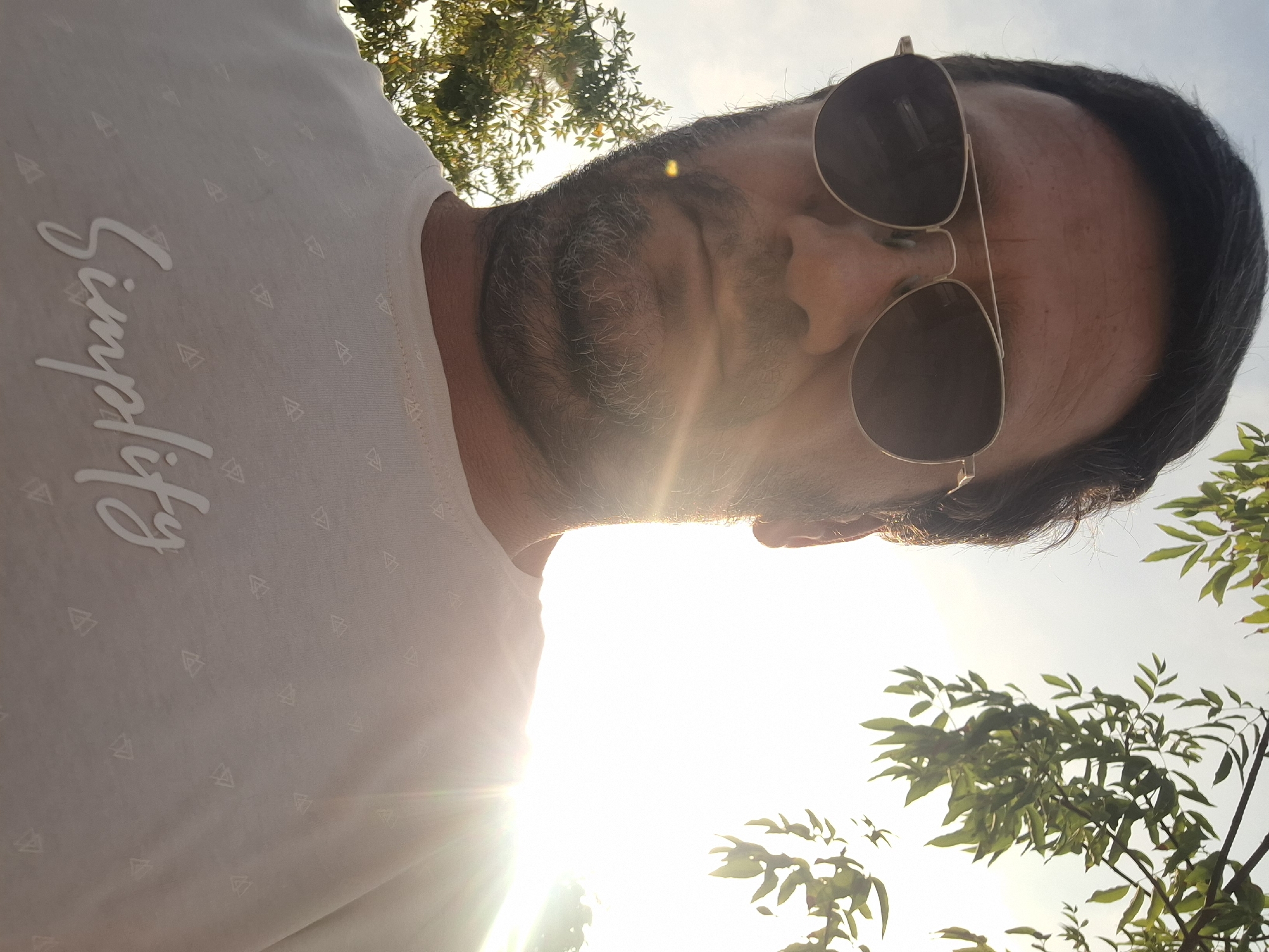 A low-angle selfie shows a man wearing aviator sunglasses and a white t-shirt with a subtle geometric pattern and the word "Simplify" printed in cursive. The man has dark hair and a beard, and is looking directly at the camera. Sunlight beams from the upper right corner of the image, creating lens flare. Green leaves from a tree are visible in the upper and lower right corners of the frame. The sky is clear and bright blue. The image appears to have been taken outdoors in Yerocham, Israel, likely during daylight hours.