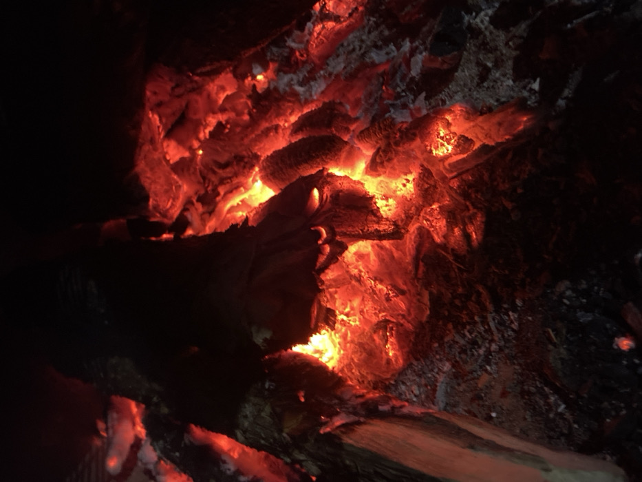 The scene depicts a close-up view of an active fire, consisting of intensely glowing red and orange embers intermixed with charred, partially consumed wood and white-grey ash. Dark, unburnt sections of wood are visible, primarily along the left and top, contrasting with the radiant heat emitted by the burning material below. The ambient environment is dark, indicating the fire is the primary light source. No individuals, identifiable structures, or external objects are present within the frame. This event occurs in the city of Los Velásquez, Venezuela.