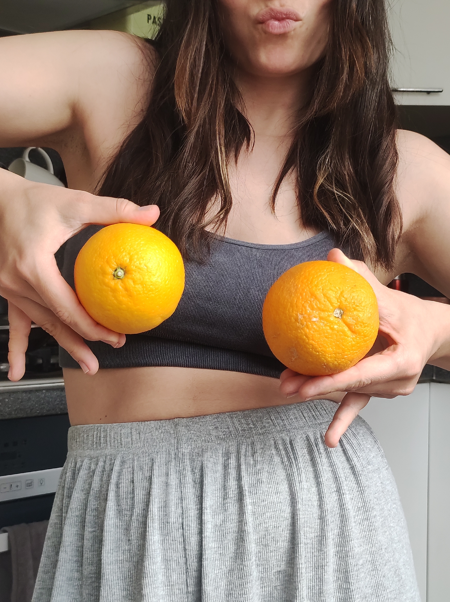 A woman with long brown hair, pursed lips, wearing a gray sports bra and a gray pleated skirt, is holding an orange in each hand in what appears to be an indoor kitchen setting. The background includes kitchen cabinets and appliances. The location of the image is not discernible from the provided information.