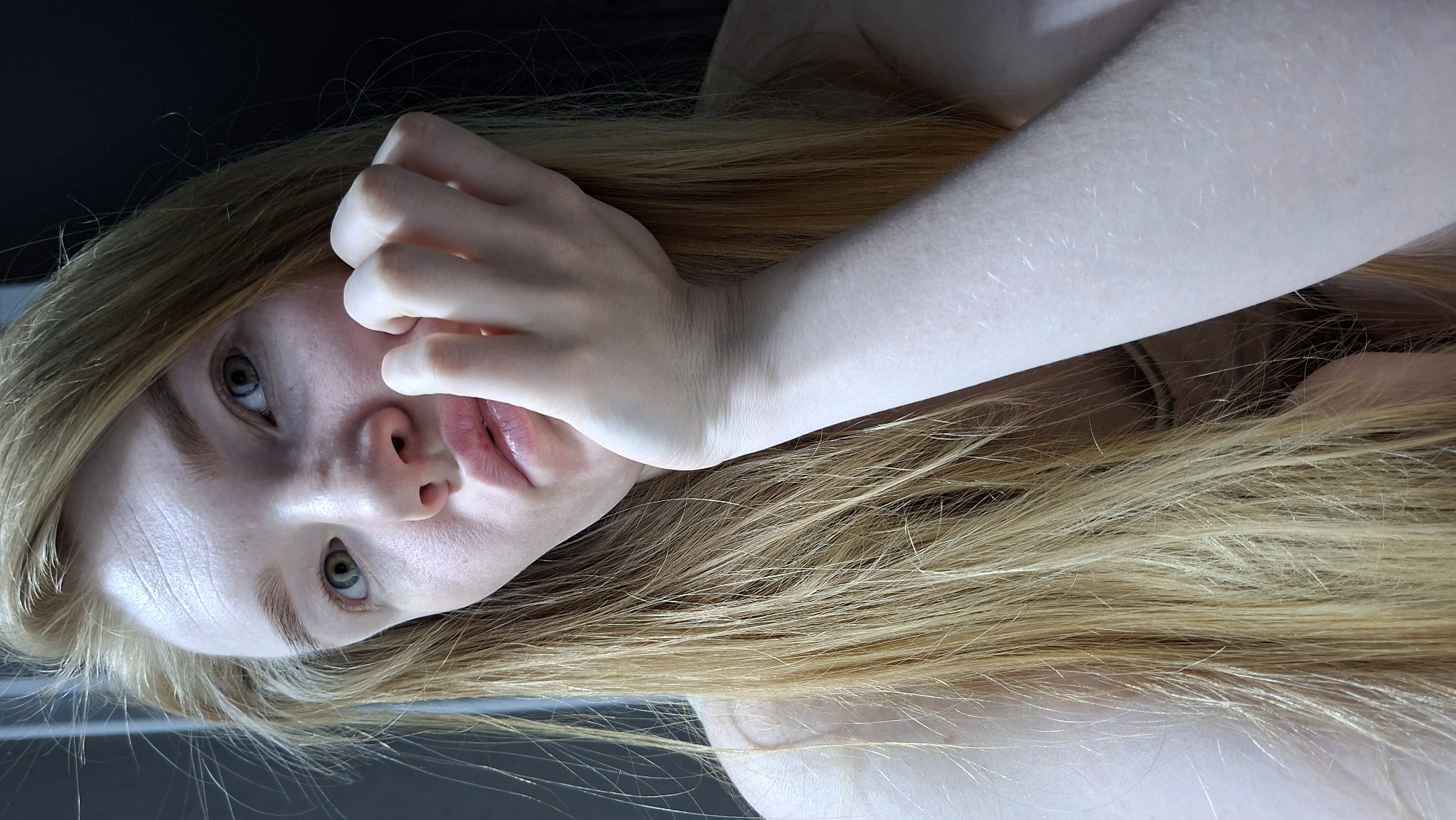 A young, light-skinned individual with long, light brown hair is depicted in a close-up, rotated image. The person's head is tilted, with their right hand near their face, fingers partially obscuring their cheek. The person's eyes are open, and their expression is neutral. The background is dark and out of focus. The location cannot be determined as Chaikovsky, Russia, based on the visual information provided. There is no identifying location.
