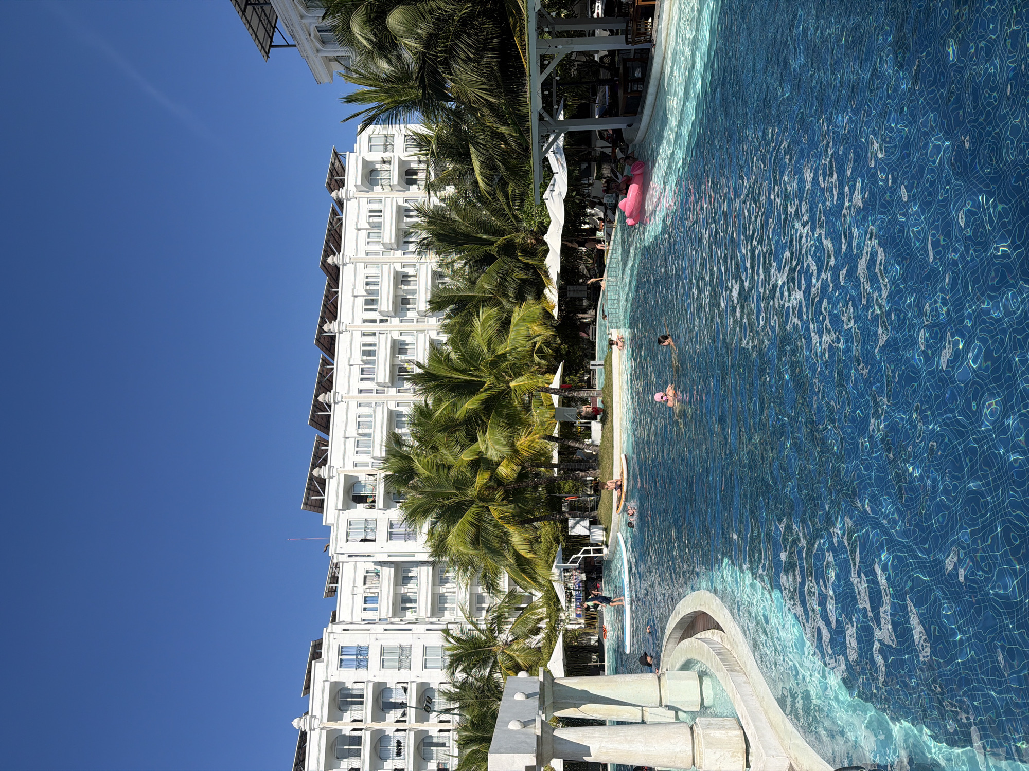 This image depicts a sun-drenched resort swimming pool in Nha Trang, Vietnam. The pool, with its clear blue water and tiled bottom, is the central focus. Several people are enjoying the water, with some swimming, others lounging on inflatable devices, and a few standing on paddleboards. In the background, a modern, multi-story white hotel building stands against a bright blue sky, its balconies adorned with potted plants. Numerous palm trees with lush green fronds are scattered around the pool area, providing shade and tropical ambiance. White umbrellas and a covered bar area are also visible, suggesting amenities for resort guests. The scene conveys a sense of relaxation and vacation.