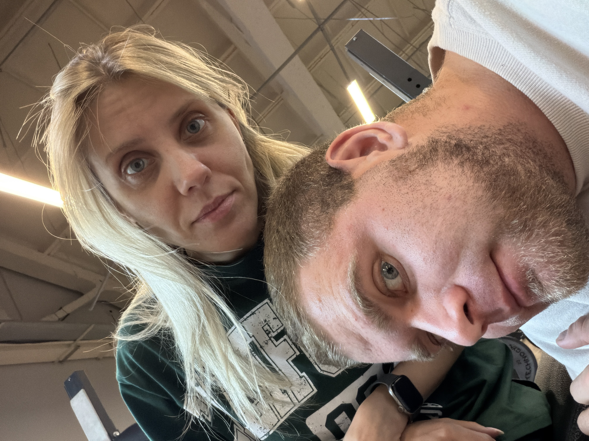 The image is a selfie taken from a low angle, featuring a man and a woman. The man is in the foreground, with his face partially visible and looking towards the camera. He has short brown hair and a beard. The woman is positioned behind him, with her face also visible and looking towards the camera. She has long blonde hair. Both individuals are wearing casual clothing. The background appears to be an indoor environment, possibly a gym, with what looks like gym equipment and ceiling lights. There are no clear indications of the specific location in Chaikovsky, Russia.