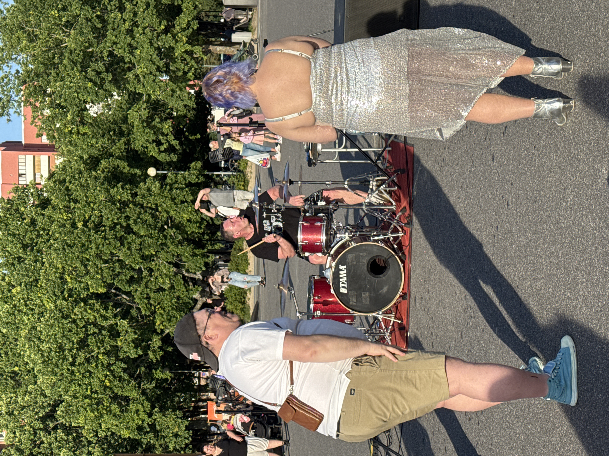 In Tallinn, Estonia, a male drummer plays a drum set on a red mat in an outdoor setting. He is wearing a black t-shirt and patterned shorts. To his left, a man in a white t-shirt, khaki shorts, and a baseball cap stands facing away from the camera, with a brown satchel slung across his chest. To the drummer's right, a person with purple-tinted hair, wearing a shimmering silver dress and silver boots, faces away from the camera. In the background, several trees line a path with a building visible beyond them. Other people and a stroller are also present in the background. The ground appears to be asphalt, and shadows suggest it is a sunny day.