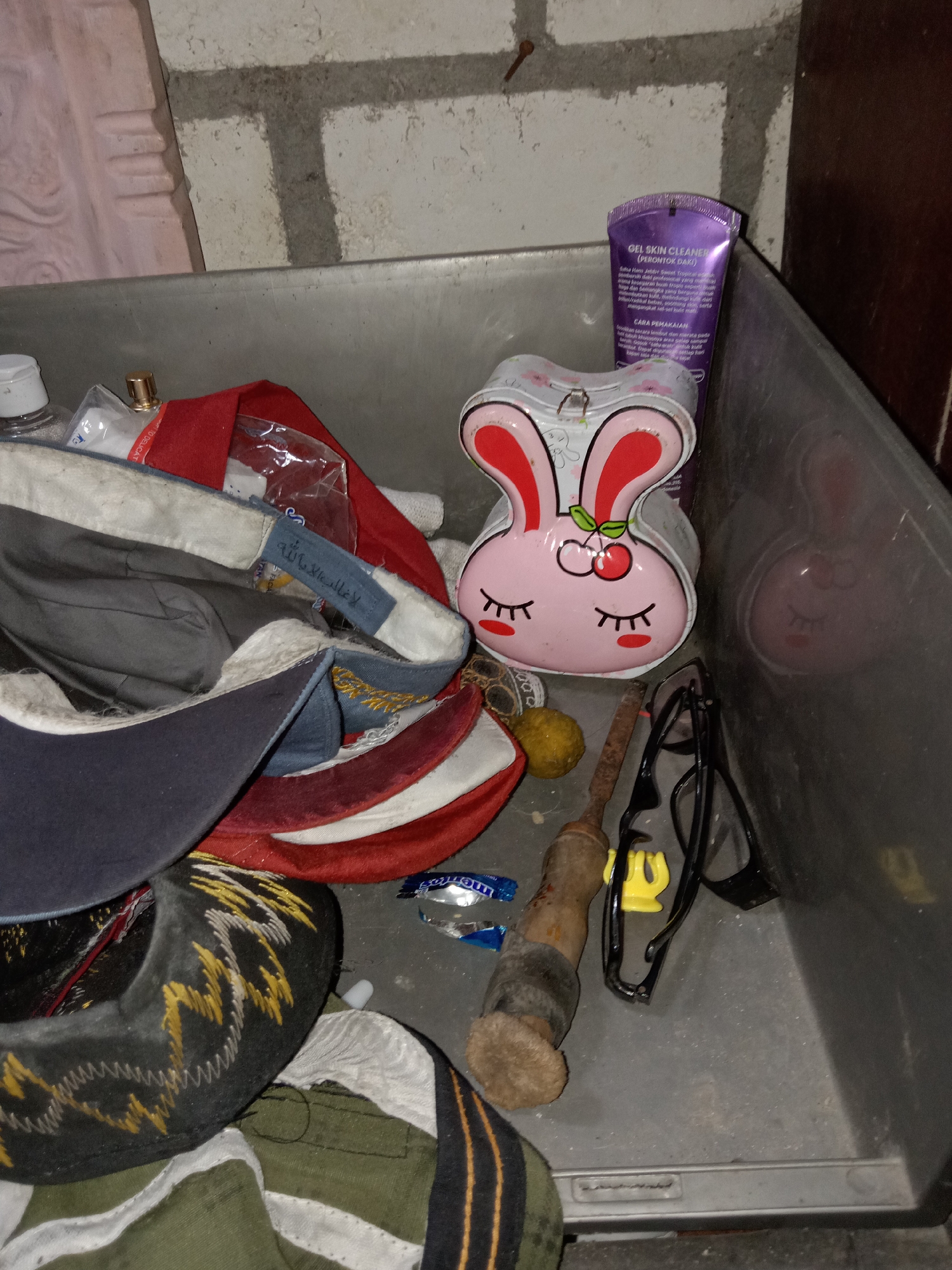The image depicts the interior of a grey plastic bin, likely a storage container, situated in a dimly lit environment. The bin contains a variety of objects, suggesting a disorganized collection. Prominently featured is a pink, bunny-shaped tin with closed eyes and cherry accents, possibly a confectionery container. Adjacent to it is a purple tube of "GEL SKIN CLEANER (PEMBERSH DAKI)" with Indonesian text, indicating a skincare product.
Several pieces of headwear are visible, including what appears to be a dark blue baseball cap with a white interior lining and a blue tag with Arabic inscription, and other caps with red and white sections. There are also other assorted items, such as a small yellow spherical object, a wooden-handled tool with a metallic end, and a pair of black-rimmed glasses. A piece of foil-wrapped candy or gum lies on the floor of the bin. The background consists of a rough, light-colored brick wall with dark grout lines. The overall impression is that of a miscellaneous collection of personal items stored in a utilitarian container. The location of Jumput, Indonesia, is not visually ascertainable from the image itself.