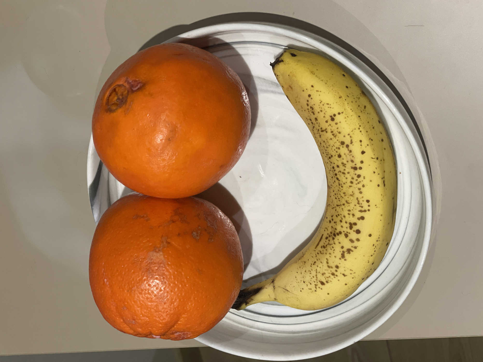 A top-down shot shows two oranges and one banana on a white plate with gray swirls. The oranges are round and orange in color. One orange has a blemish on its surface. The banana is yellow with brown spots and curves along the right side of the plate. The plate is resting on a light beige surface. There is no indication of the location being Batumi, Georgia.