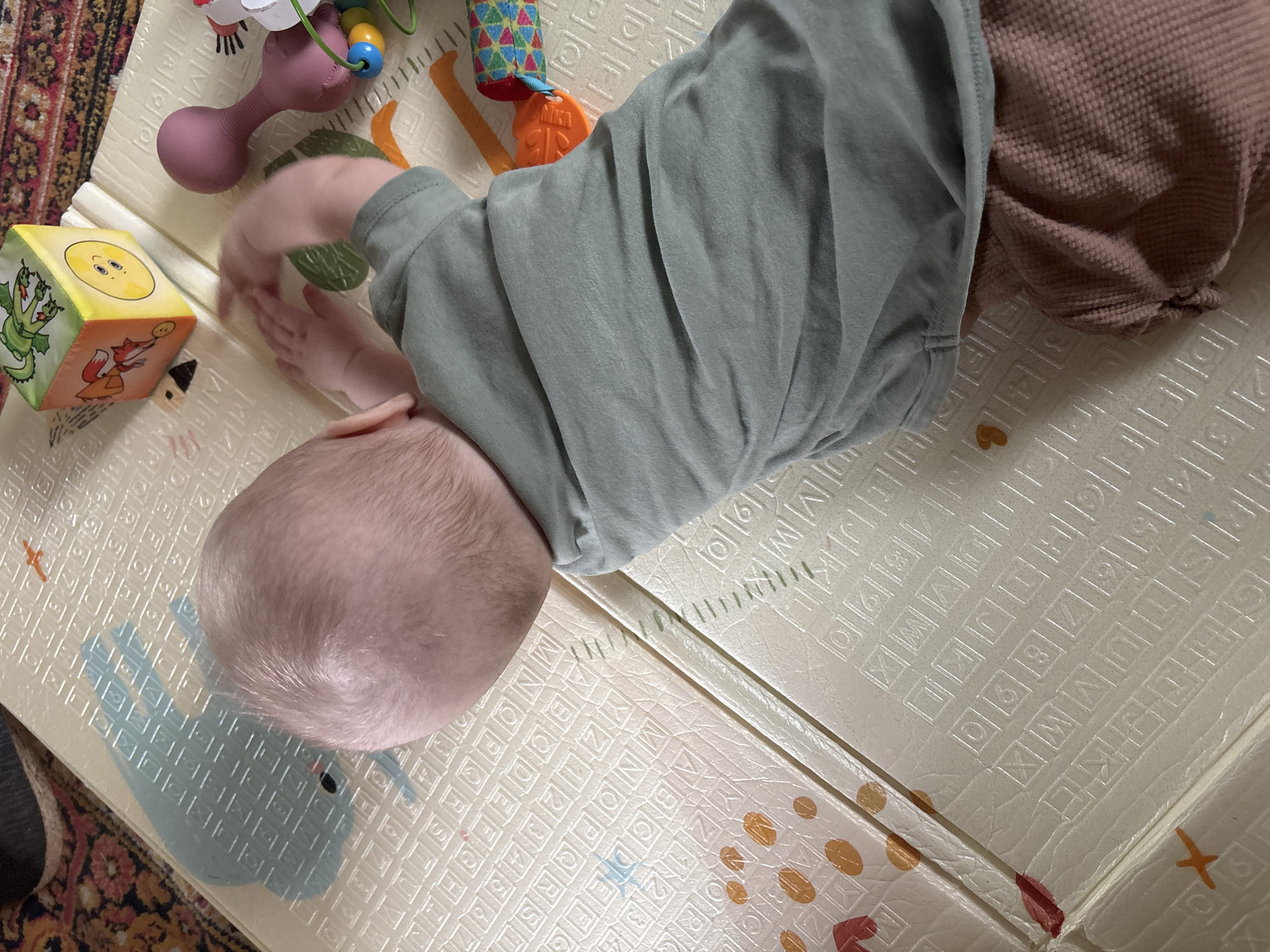 A baby is lying on an alphabet foam play mat. The baby has a light-colored head of hair and is wearing a light green long-sleeved shirt and mauve-colored pants. To the baby's left are several toys, including a plush block with images of animals and a pink toy with colored beads. The mat is patterned with letters and numbers. A portion of a patterned rug is visible in the lower left corner. The location in the city of Мещёры, Russia is not discernible from the provided image.