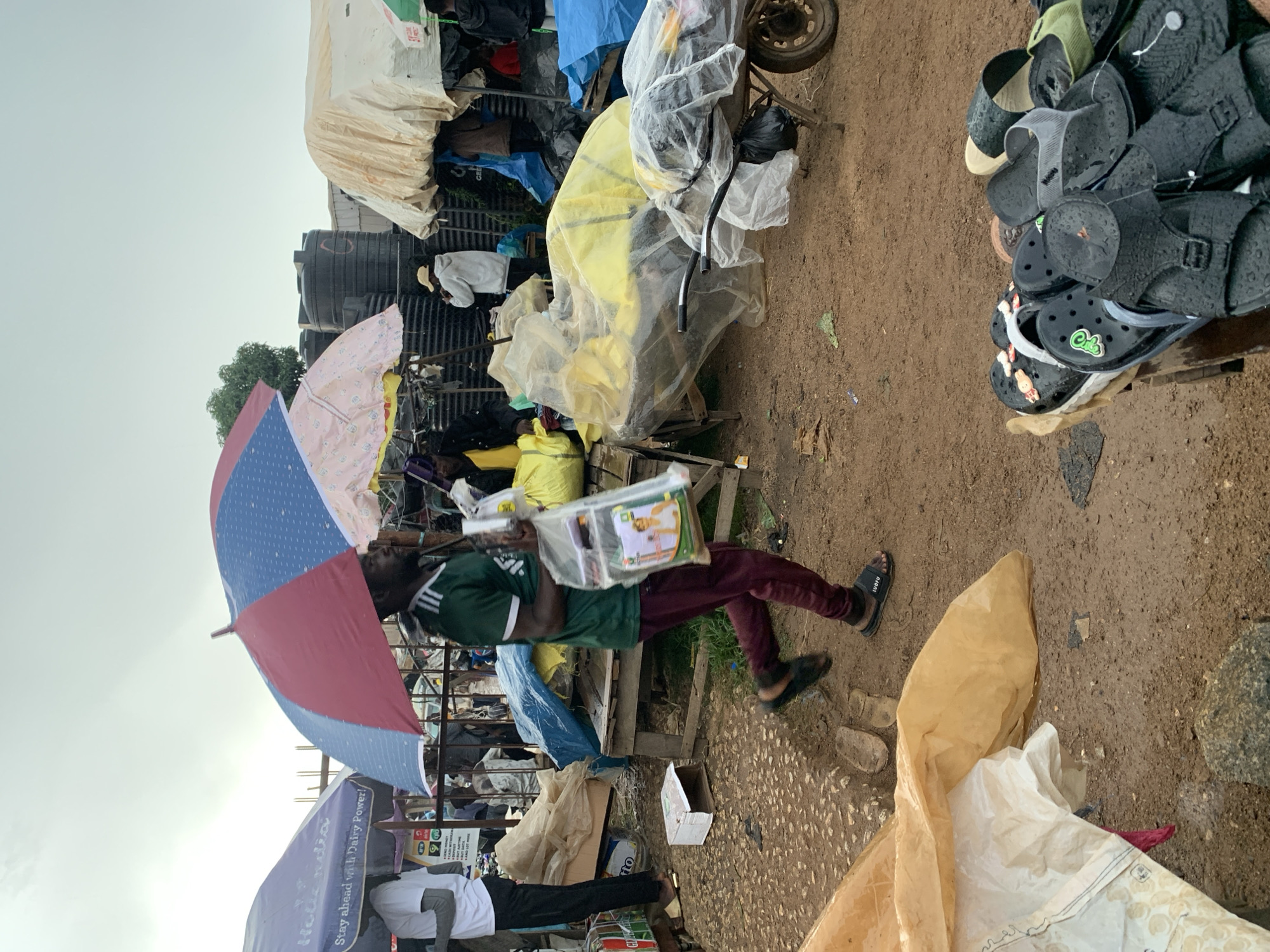 The image depicts a bustling market scene in Gura Top, Nigeria. In the foreground, a man wearing a green t-shirt and maroon pants walks briskly under a red and blue umbrella, carrying a plastic bag filled with items. He is wearing black sandals. The ground is muddy, indicating recent rain. To the left, another person in a white t-shirt and black pants is partially visible under a purple umbrella. Various stalls with tarps and umbrellas are set up, displaying goods covered in plastic sheeting. In the background, large black water tanks are visible, along with more market stalls and structures. A wheelbarrow covered in plastic is positioned to the right. A pile of sandals is prominently displayed in the lower right corner of the frame. The overall atmosphere suggests a busy, open-air market environment during overcast weather.