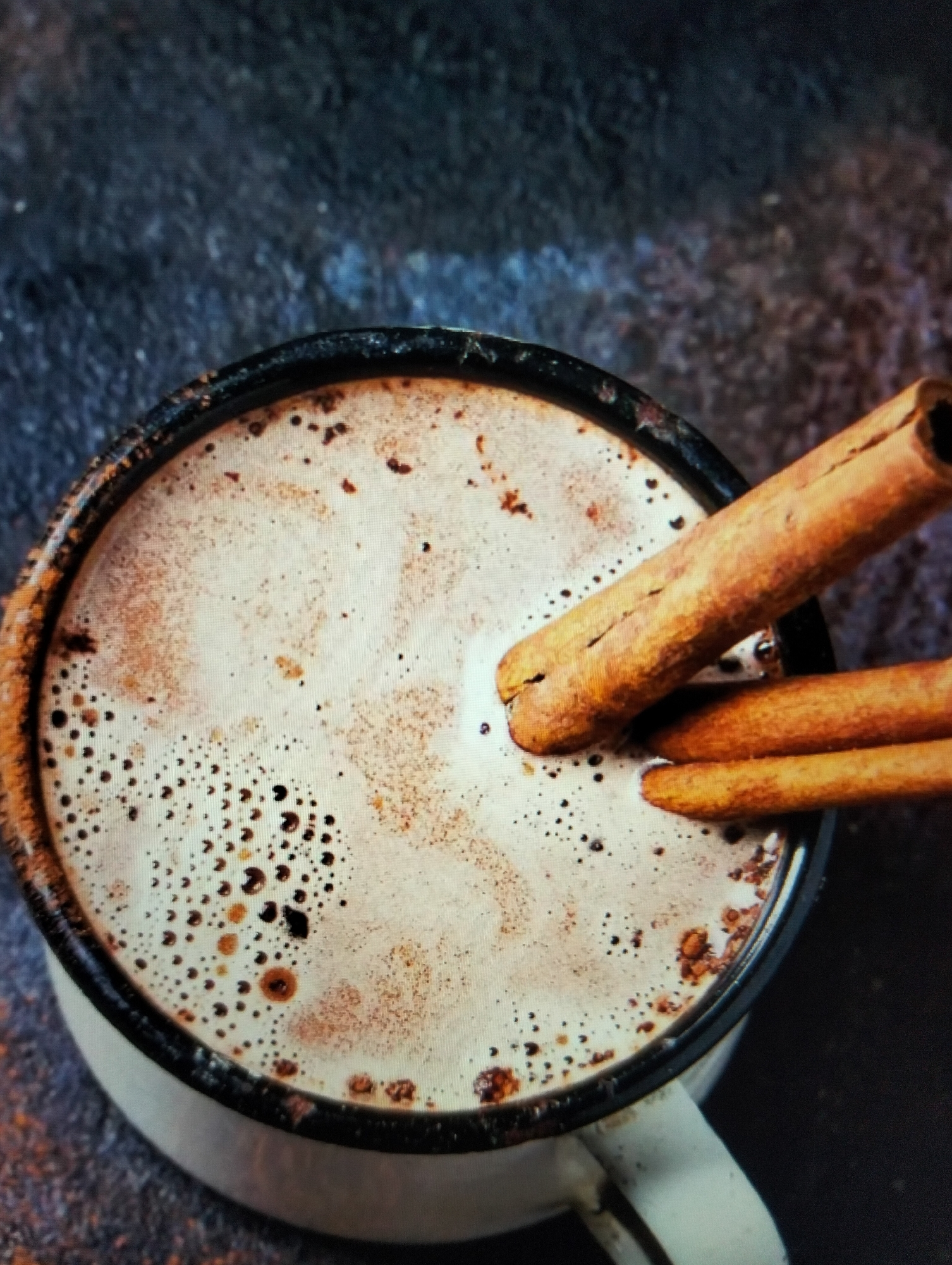 The image is a close-up shot of a mug of hot chocolate with cinnamon sticks. The mug is white with a black rim and is filled with a frothy, light brown liquid. Two cinnamon sticks are placed in the mug, with one extending out of the top. The background is a dark, textured surface. There are no people or any indications of location in the city of Payagaboh, Indonesia.