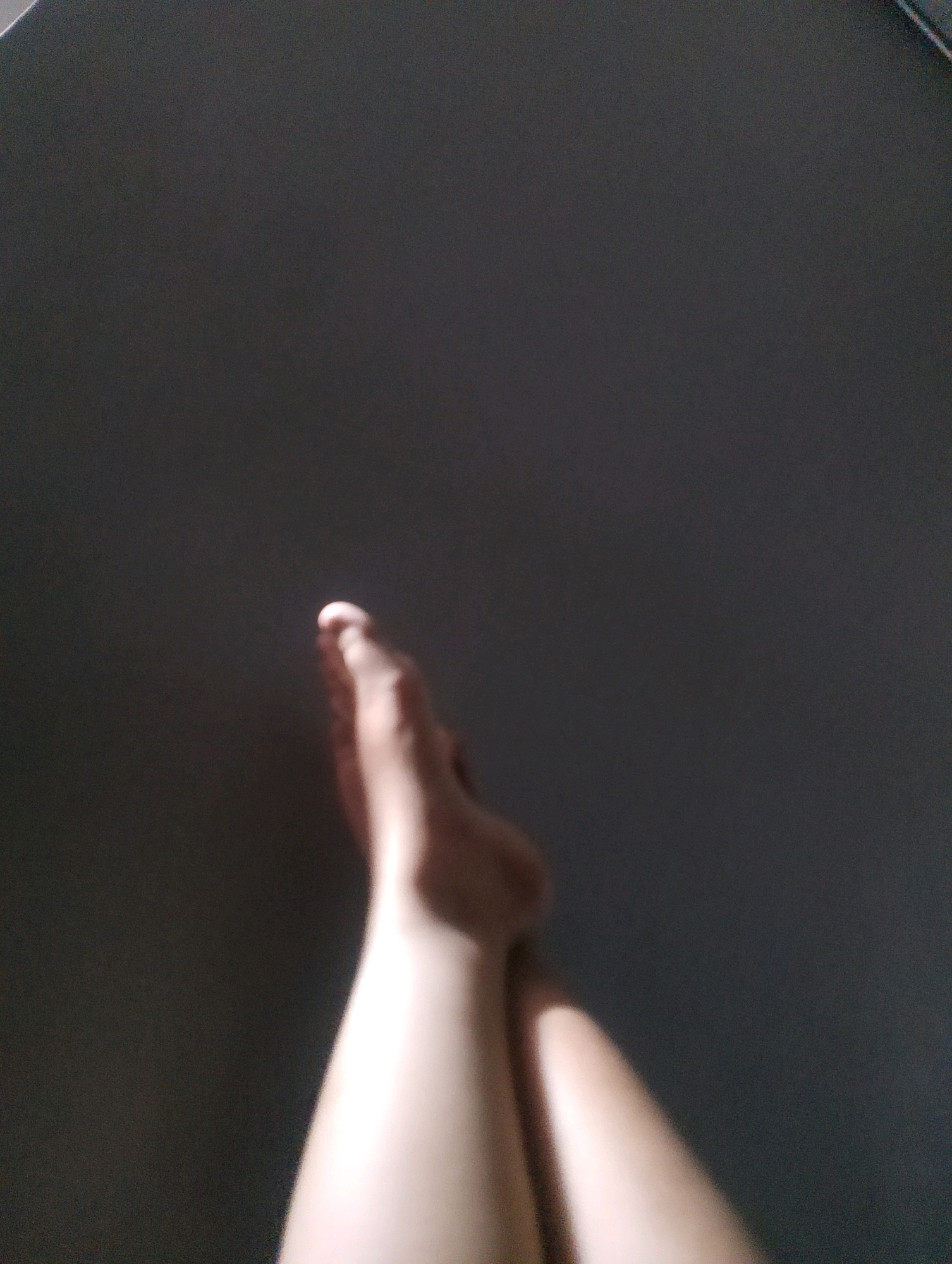 The image shows two legs and feet of a person lying down. The background is a dark, solid color, possibly a wall or floor. The lighting suggests a natural light source coming from the left, casting shadows and highlights on the legs. There is no discernible urban environment or specific location in Magelang, Indonesia visible in the image. The scene is static, with no action or interaction depicted.