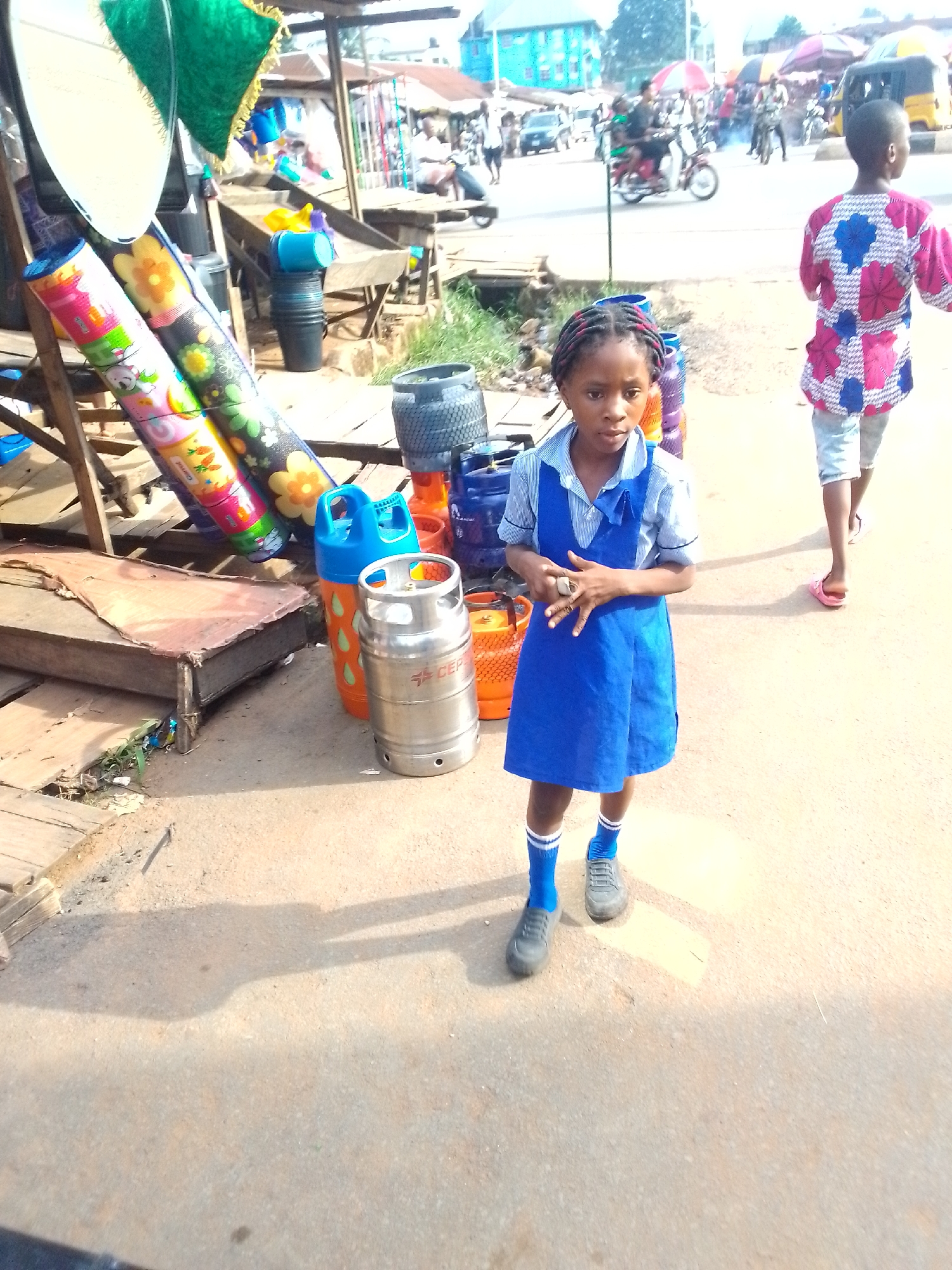 The image, taken in Orlu, Nigeria, shows a young girl in a blue school uniform standing on a paved surface, likely a street or sidewalk. She wears gray shoes and blue socks with white stripes. Her hair is braided with blue ribbons. To her left are rolled-up mats with colorful patterns, along with blue and orange gas canisters and a silver, possibly stainless steel, keg or tank. Behind her, a person in a pink and blue patterned shirt walks away. The background includes a busy street scene with people, motorcycles, cars, and open-air stalls or shops. Buildings are visible in the distance. The lighting suggests it is daytime, and there is strong sunlight casting shadows on the ground. The scene appears to be a commercial area with a mix of pedestrian and vehicular traffic.
