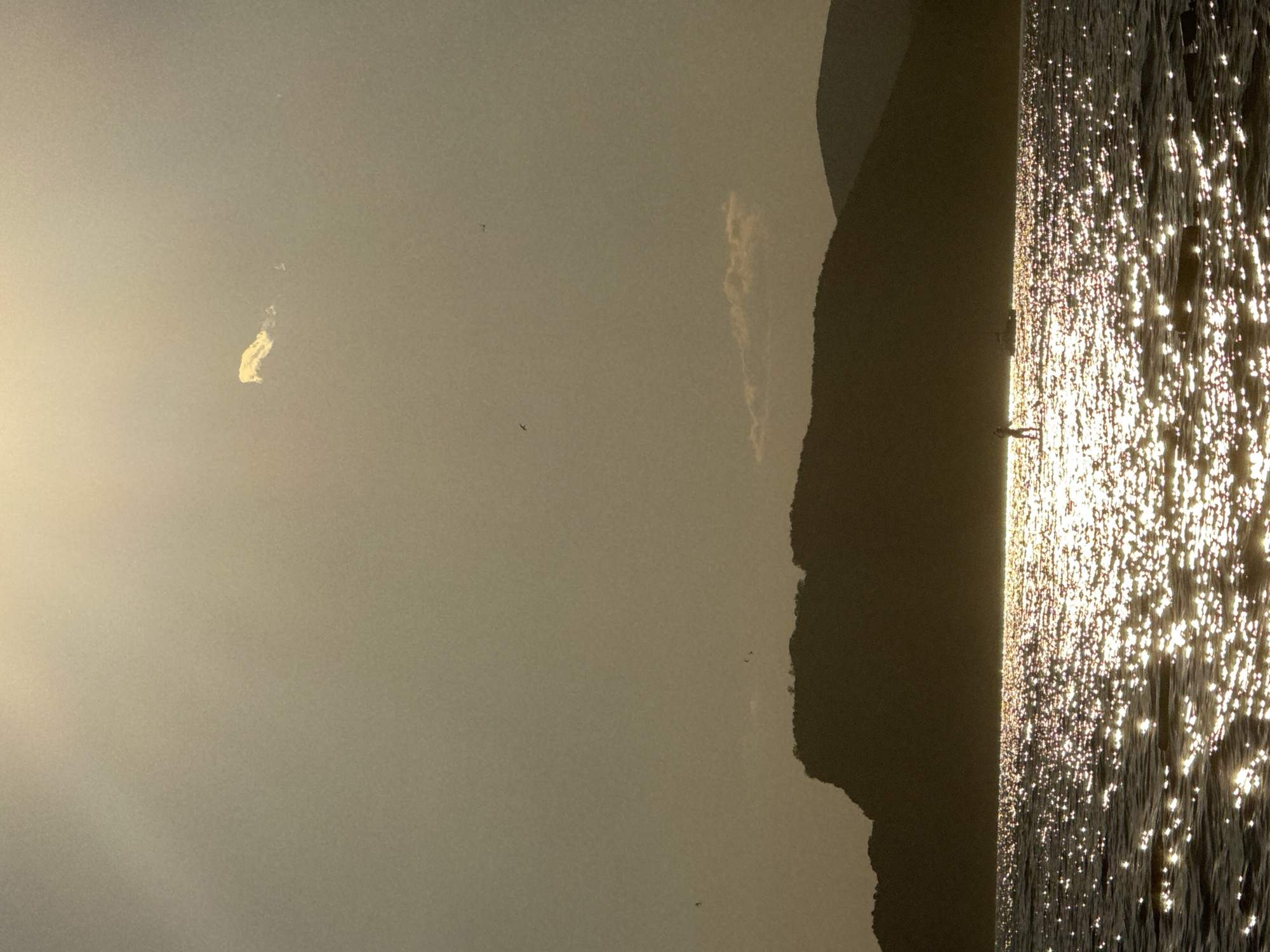 A silhouetted figure stands on a paddleboard in the sparkling water of the Adriatic Sea, with the distinctive silhouette of the Pržno coastline and hills forming the background. The sun, low in the sky, casts a strong glare on the water, creating a shimmering effect. A small, bright cloud is visible in the hazy, golden sky above. Several birds are in flight in the upper portion of the image.