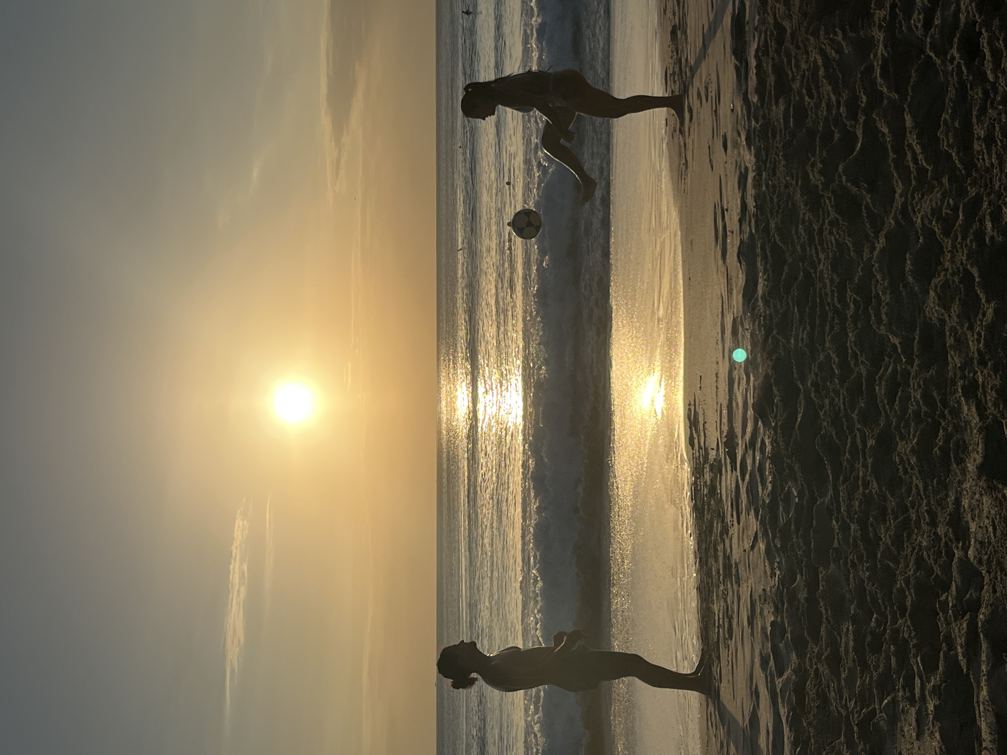 The image depicts a sunset scene on a beach in Kuta Selatan, Indonesia. Two silhouetted figures, a male and a female, are standing on the wet sand near the water's edge. The male, positioned on the left, faces the ocean with his left side visible. The female, on the right, is kicking a soccer ball with her right leg, her body angled towards the male. The sun is setting over the ocean, casting a golden glow across the sky and reflecting on the water's surface. Waves are breaking on the shore, and the sand in the foreground is textured with footprints and ripples.