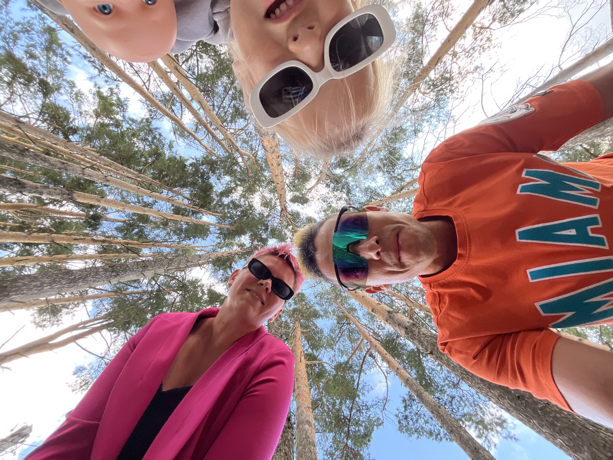 A low-angle, wide-shot photograph captures three individuals seemingly posing for a selfie outdoors amidst a forest of tall, thin trees. The perspective is from below, looking up towards the sky and the faces of the subjects. In the foreground, a man wearing an orange t-shirt with the word "MIAMI" in turquoise letters and tinted sports sunglasses looks directly into the camera. To his left, another individual wearing a bright pink jacket and black sunglasses, with pink-dyed hair, is also looking towards the camera with a slight smile. To the right and slightly above, a child with blonde hair and large white sunglasses is visible, their face partially obscured. The sky above is a bright blue with scattered clouds, and the canopy of pine trees creates a natural, dappled lighting effect. The image appears to be taken in a natural, wooded area, possibly a park or forest trail, and the casual attire suggests an informal outdoor activity. The specific location in Chaikovsky, Russia, is not identifiable from visual cues in the image alone.