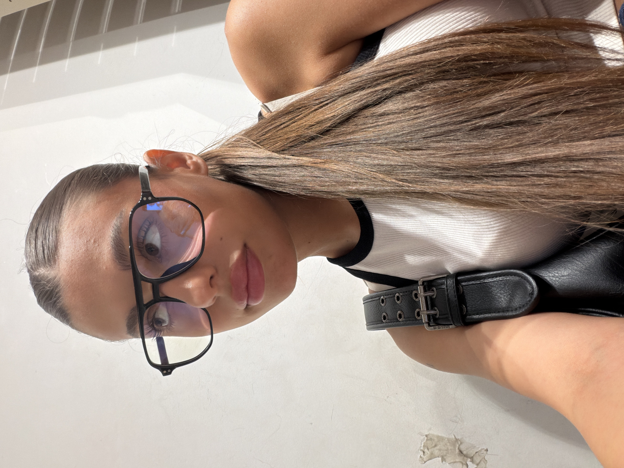 The media file shows a young woman from the chest up, oriented sideways. She has long, light brown hair pulled back, and is wearing a white ribbed tank top with a black trim. She is also wearing black-rimmed glasses and a black belt with a silver buckle and two rows of eyelets. The background is a plain white wall, with shadows cast on it from what appears to be blinds or a window. The lighting suggests it is daytime. The woman is looking directly at the camera, with a neutral expression. The image was captured in Burgas, Bulgaria.