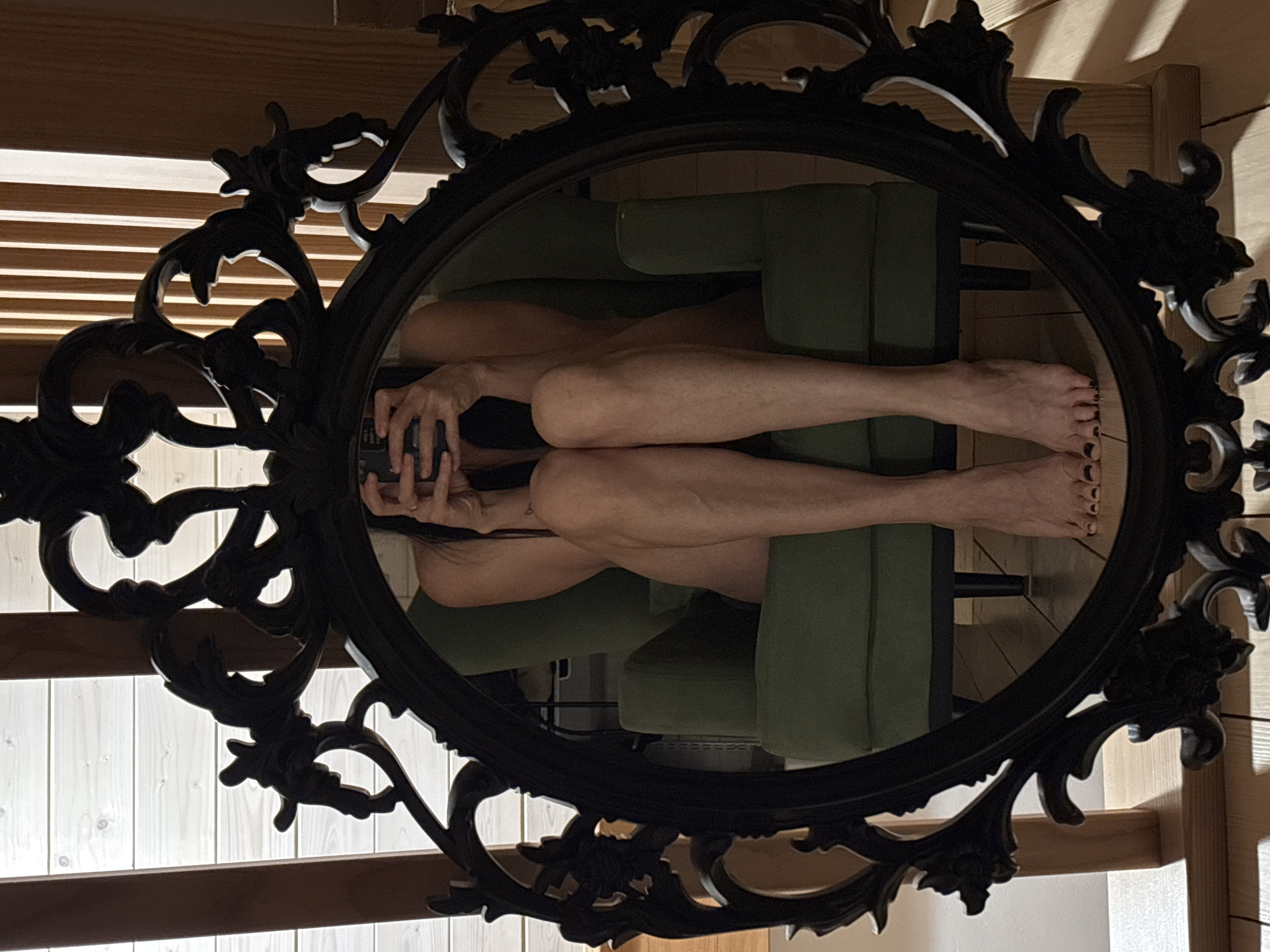 A reflection in an ornate black oval mirror shows a person lying on an olive green sofa. The person's legs are visible, and they are holding a dark-colored object, possibly a phone, in their hands. The background visible through the mirror includes wooden blinds and portions of a wooden structure. The location is not discernible from the image to the level of specificity of a city such as Chaikovsky, Russia.