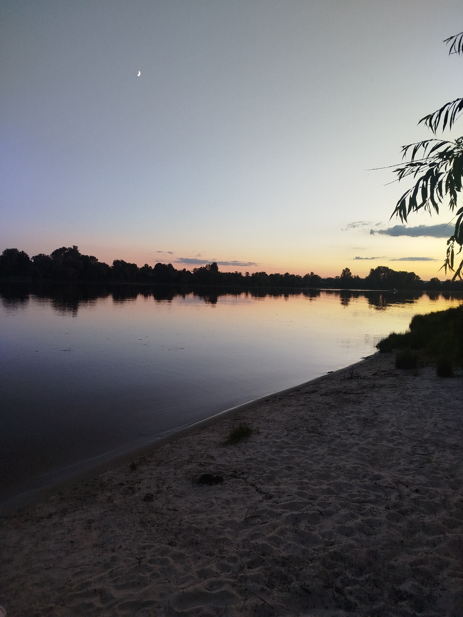 The image depicts a serene twilight scene by a body of water. A crescent moon is visible in the upper left quadrant of the sky, which transitions from a pale blue to hues of orange and yellow near the horizon. The sky is mostly clear, with a few wispy clouds. A silhouetted treeline runs along the far bank of the river, its reflection shimmering on the calm water. The foreground consists of a sandy beach with some sparse vegetation. To the upper right, a dark, leafy branch of a tree hangs into the frame. The overall impression is one of tranquility at dusk. The image does not contain any people or provide any specific information about the location within the city of Unknown.
