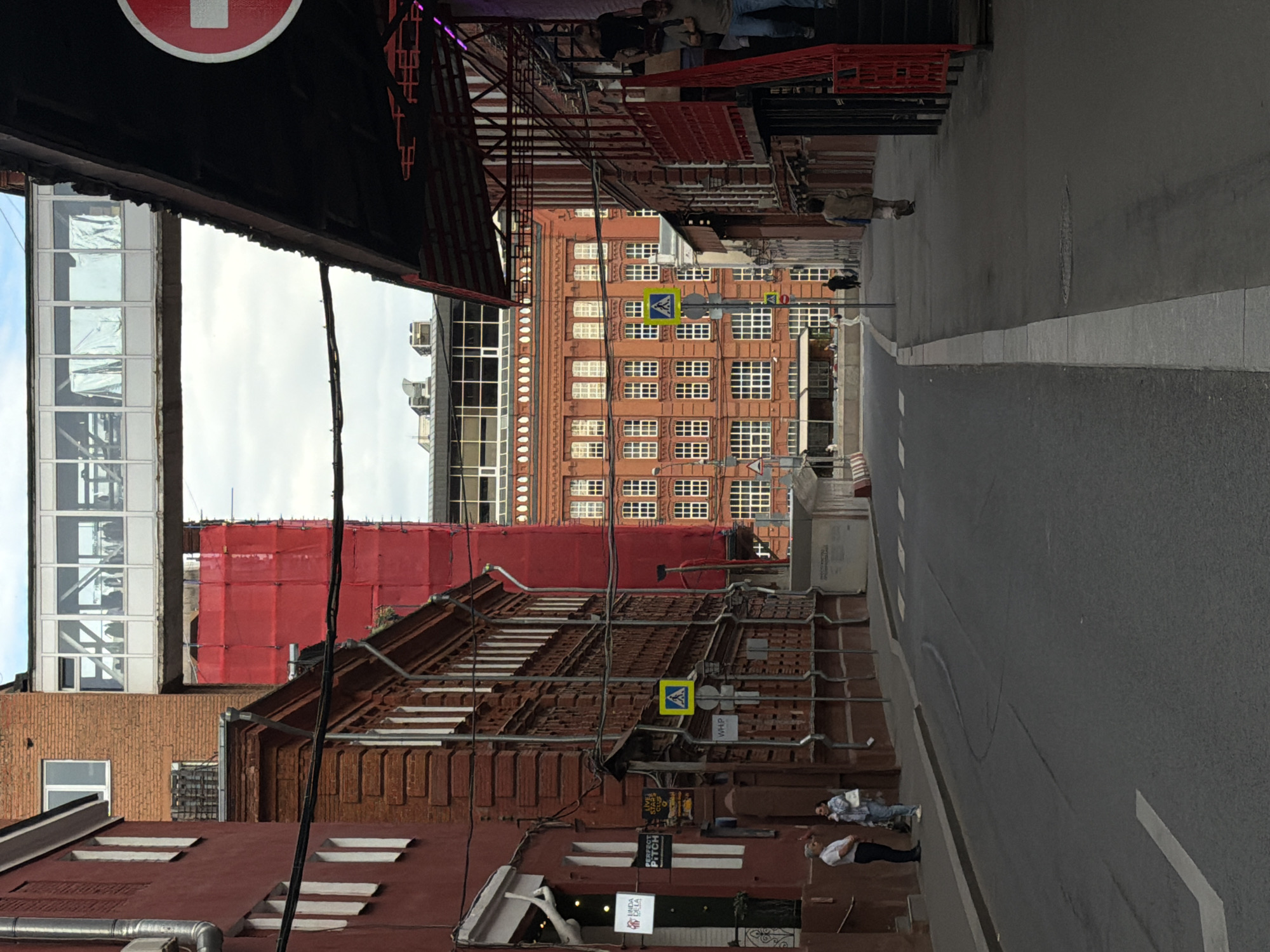The image depicts a street scene in an urban environment, likely a city. The buildings on either side of the street are constructed primarily of red brick. Scaffolding and possibly construction netting are visible on the lower buildings, indicating ongoing construction or renovation work.
Two pedestrians are visible on the right side of the street, close to one of the brick buildings. The street itself is paved and includes painted lane markings. There are crosswalk signs on the streets.
The style of architecture and the presence of construction work suggest a historic urban area potentially undergoing modernization or restoration. The location within the city and country remains unknown based solely on the provided image.