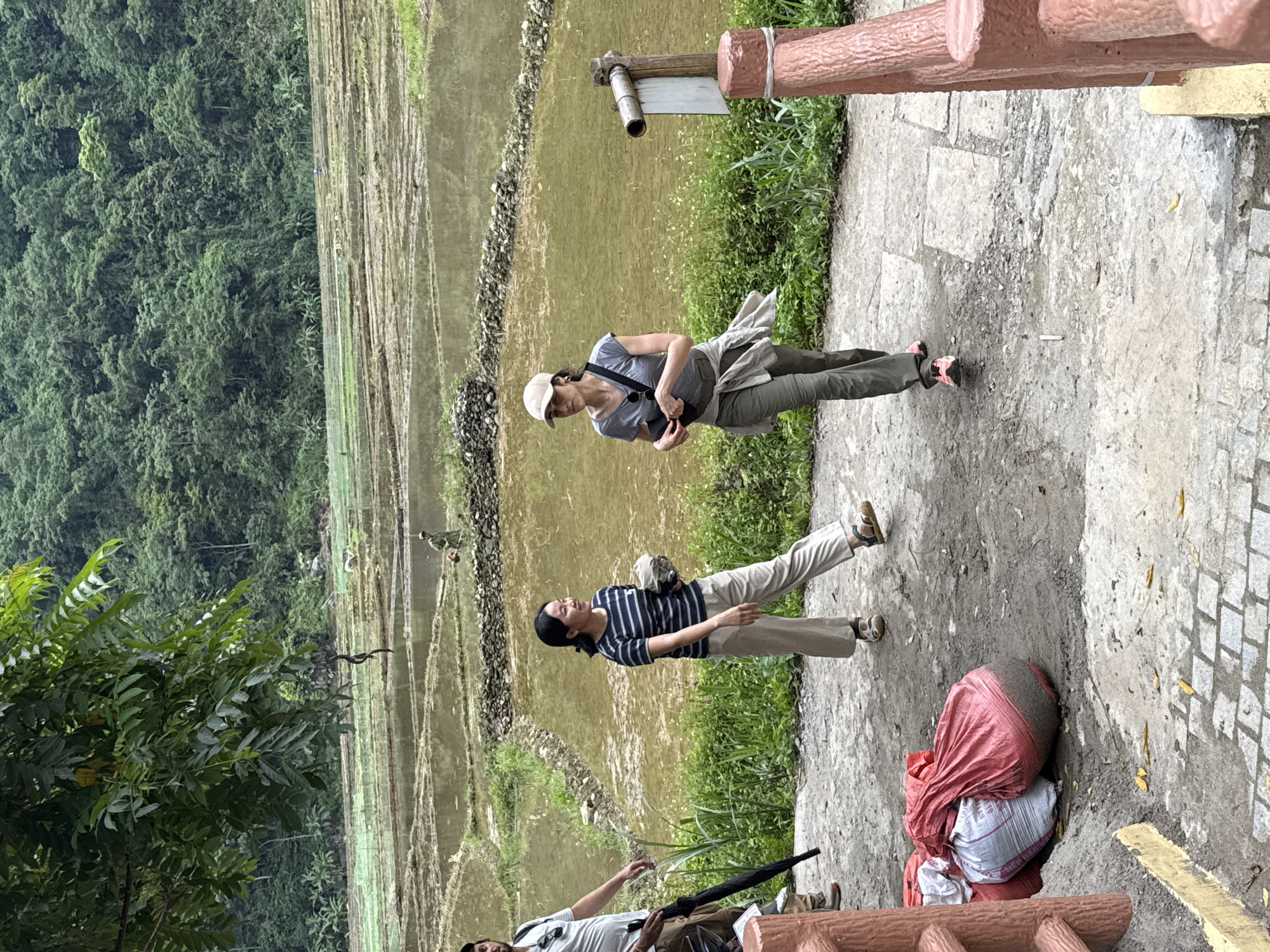 In Pù Luông Commune, Vietnam, two women are walking on a paved path with lush green rice terraces and dense jungle foliage in the background. The woman on the left, wearing a striped t-shirt and beige pants, carries a light-colored object in her hand. The woman on the right, dressed in a grey t-shirt, olive-green pants, and a beige cap, has a dark bag slung across her body and another bag around her waist. A third person, a man in a white t-shirt, is partially visible on the left, holding a black umbrella. In the foreground, a pile of red and white sacks filled with dark material rests on the ground. The rice terraces are flooded with water, reflecting the overcast sky.
