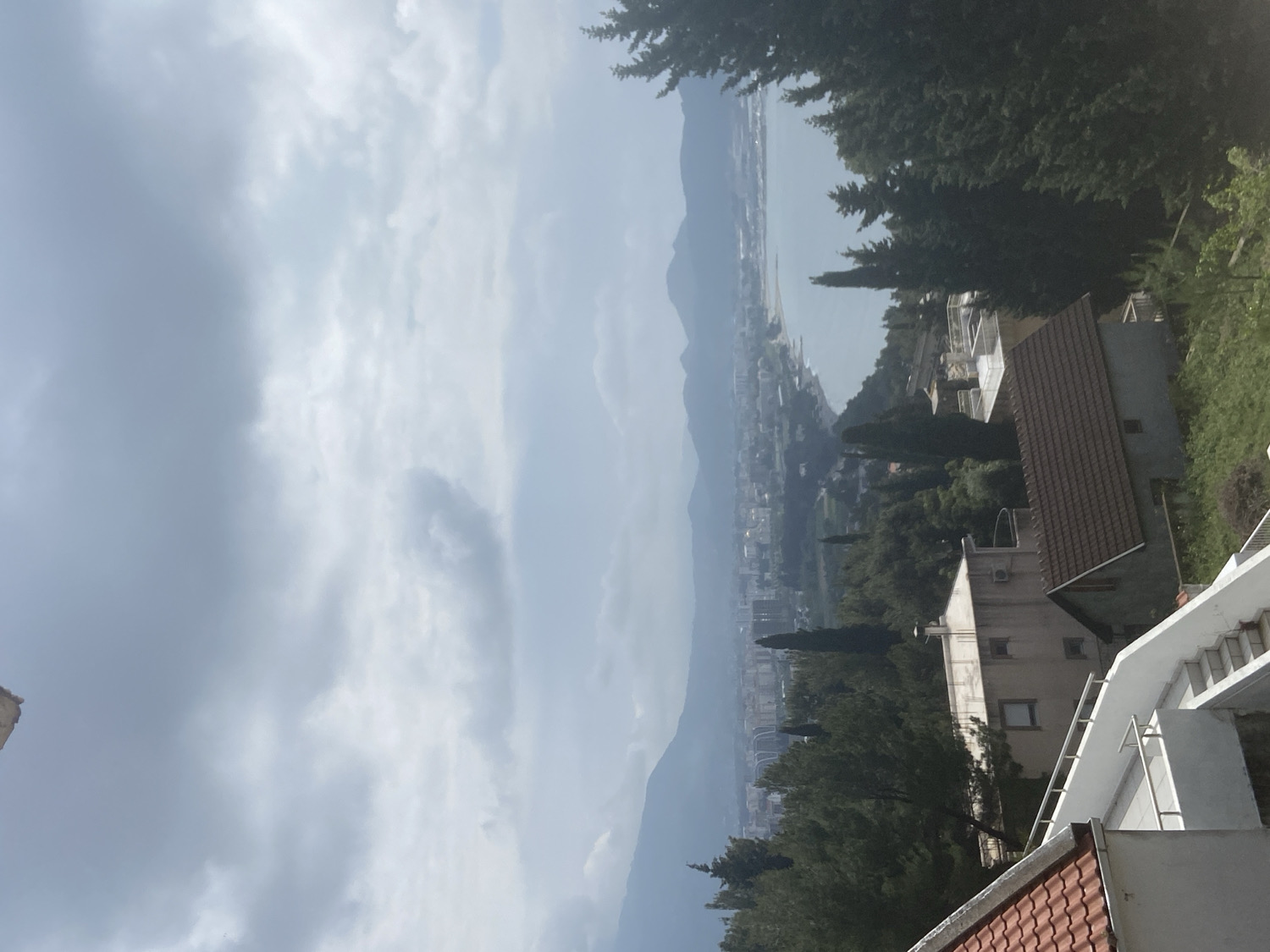 An expansive, panoramic view captures a coastal landscape from an elevated perspective, likely a hillside or mountain slope in Šušanj, Montenegro. The scene is dominated by a sweeping vista of a town or city along the shoreline, bordered by mountains and the sea, all under an overcast sky.
In the foreground, on the right side, elements of several buildings are visible. A white building with a prominent outdoor staircase and metal railings leads downwards, suggesting terraced architecture. Next to it, a corner of a roof with reddish-brown tiles can be seen. Scattered further into the midground are more residential structures, characterized by tiled roofs and light-colored walls, interspersed with lush green trees, including tall conifers and other deciduous foliage.
The midground reveals the main settlement, a coastal town stretching along the edge of a calm body of water, which is likely the Adriatic Sea. The town comprises numerous buildings nestled among abundant greenery. Beyond the town and the water, a long chain of mountains forms the background, their peaks and slopes appearing muted and somewhat hazy under the atmospheric conditions.
The sky, occupying the upper left of the image, is heavily overcast with layers of grey and white clouds, indicating a cloudy daytime. The lighting is soft and diffused, creating a serene and somewhat subdued ambiance. No people or specific activities are visible within the frame, and there is no discernible text. The scene conveys a sense of tranquility and the natural beauty of a mountainous coastal region.