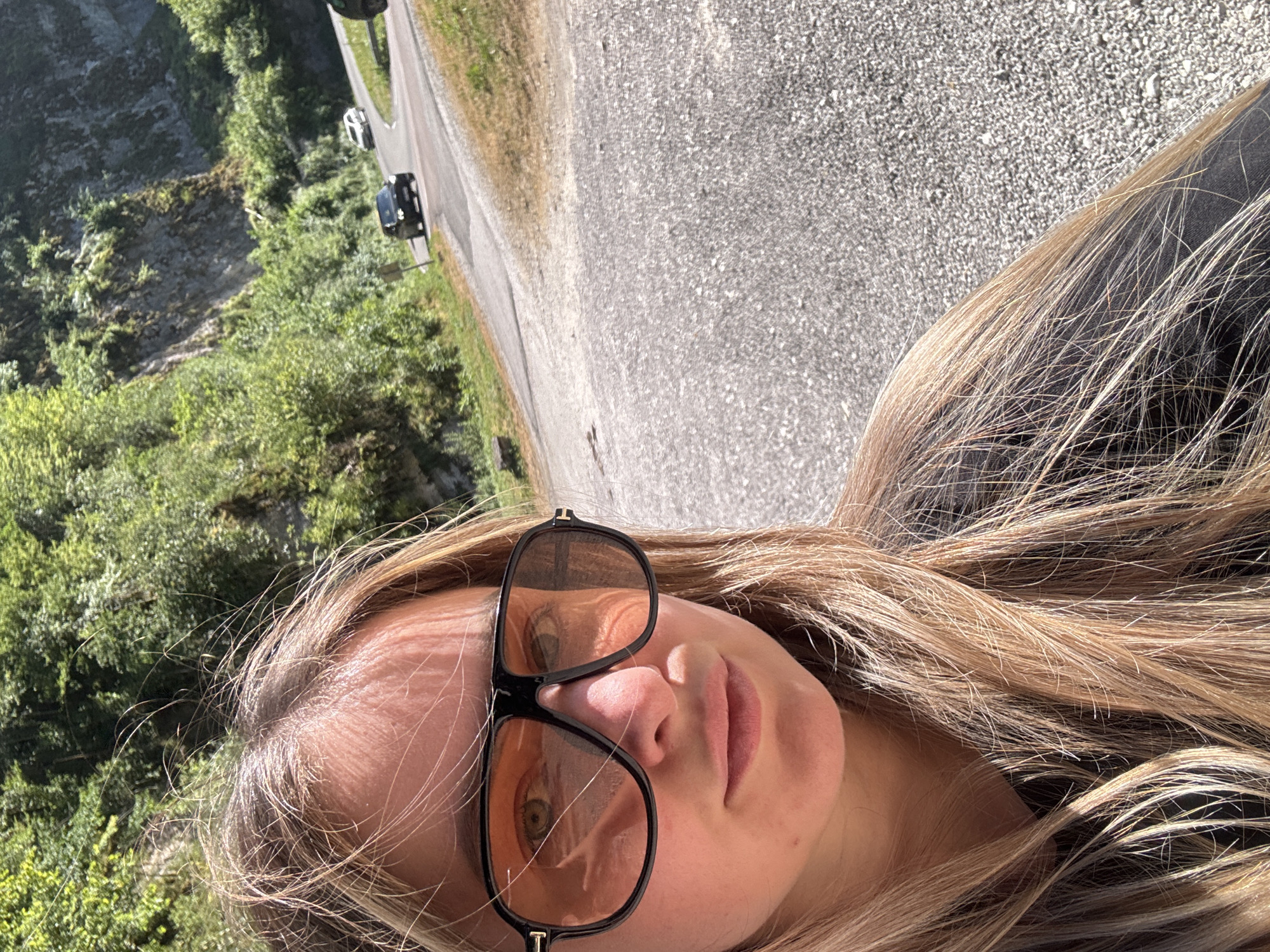 A close-up, selfie-style portrait of a young woman with blonde hair and large, orange-tinted sunglasses is shown. She is positioned on the left side of the frame, her face taking up a significant portion of the foreground. Behind her, a winding asphalt road recedes into the distance, bordered by steep, wooded hillsides. Several cars are visible on the road, appearing to be in motion. The environment suggests a sunny day in a rural or mountainous area, possibly a scenic route in L'Essard, France, given the presence of the road and natural landscape. The woman's expression is neutral.