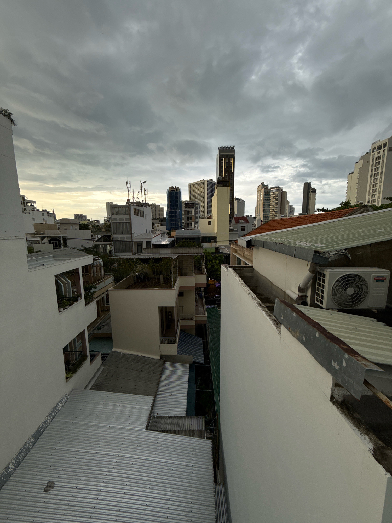 The image captures an urban skyline in Nha Trang, Vietnam, under a heavily overcast sky. The foreground is dominated by the white walls and corrugated metal roofs of residential buildings, suggesting a densely packed neighborhood. Several structures feature balconies with plants, and some have exposed wiring or makeshift additions. In the mid-ground, more buildings of varying heights are visible, some with intricate rooftop antennas and air conditioning units. The background reveals a cluster of modern, taller buildings, including high-rise apartments and commercial structures, some of which appear to be under construction or renovation. The overall impression is one of a developing city with a mix of older and newer architecture, bathed in the diffused light of a cloudy day. There are no people visible in the frame.