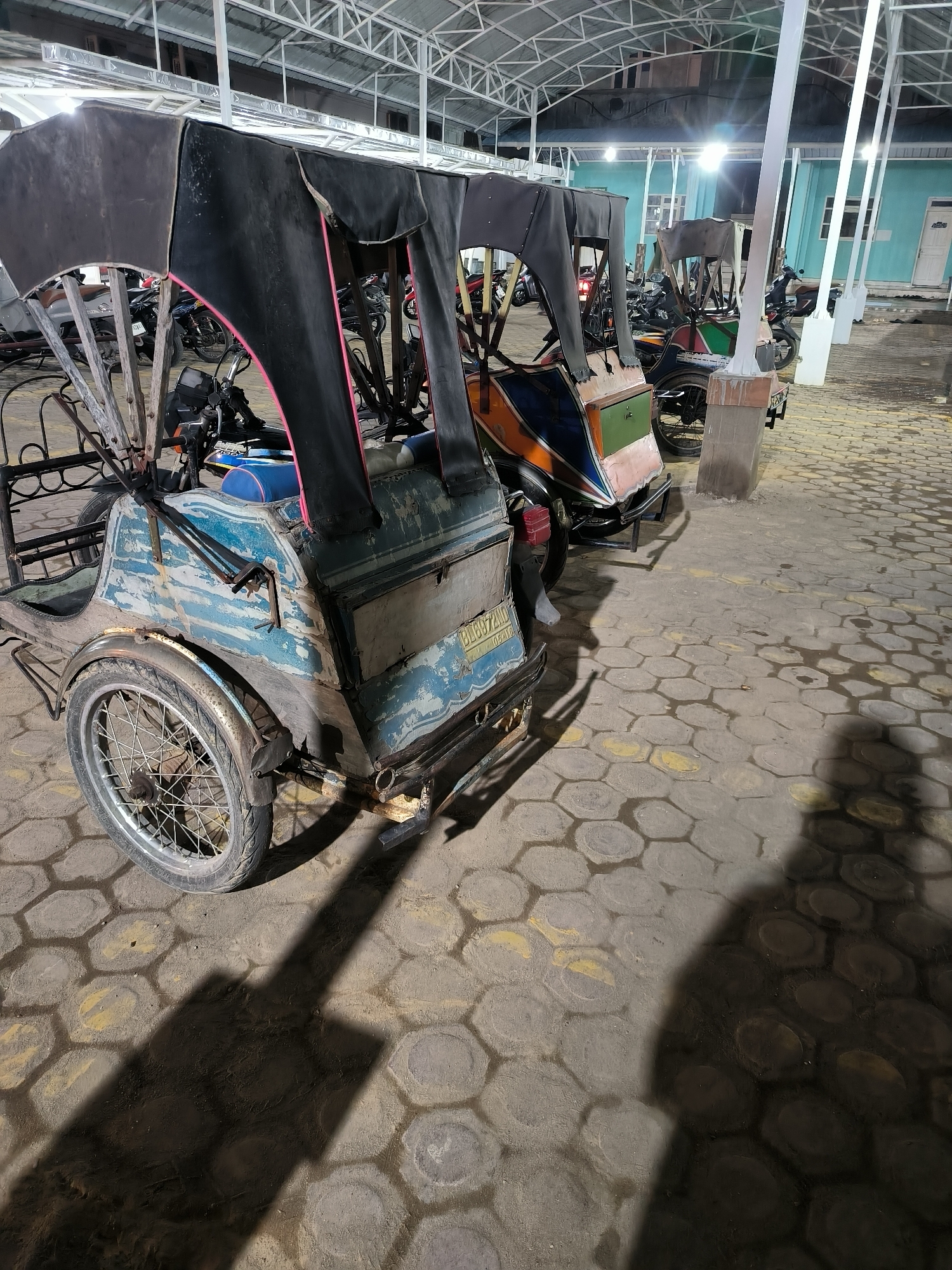 The image shows a row of parked tricycles (becaks) under a covered structure in Lhokseumawe, Indonesia. The foreground features a weathered, light blue becak with a black canvas cover. Behind it are other becaks with similar designs. The ground is paved with hexagonal blocks, and shadows are cast across the pavement. In the background, there are more vehicles, likely motorcycles and other becaks, parked near a light blue building. The scene is illuminated by overhead lighting, suggesting it is night. No people are visible.