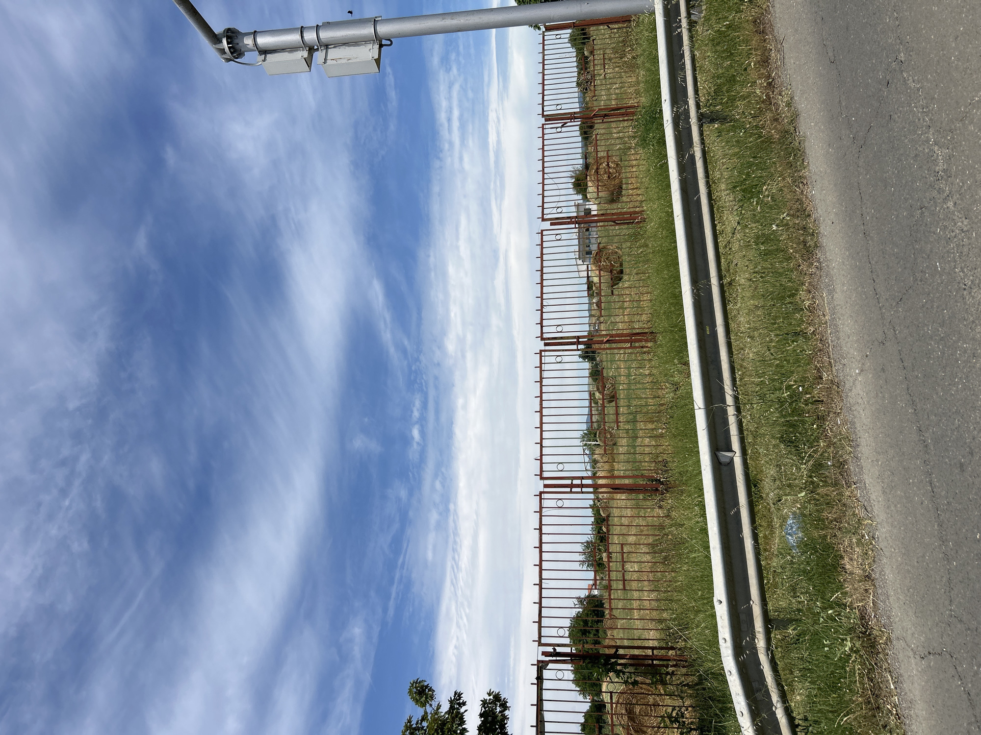 The image shows a rural roadside scene in Aheloy, Bulgaria, under a bright, cloudy sky. In the foreground, a gray asphalt road is visible with a metallic guardrail running along its edge. Beyond the guardrail is a grassy area with scattered dried stalks and some green vegetation.
A red metal fence encloses a field containing several round hay bales, some appearing dry and weathered. The fence has vertical bars and decorative circular elements. To the left, a portion of a tree with green leaves is visible. To the right, a silver pole with attached electronic boxes and an overhead arm extends into the frame, likely a street light or communication tower. The background shows more fields, some trees, and a distant building. A small bird is visible in flight near the pole. The overall impression is one of a sunny day in a semi-agricultural area.