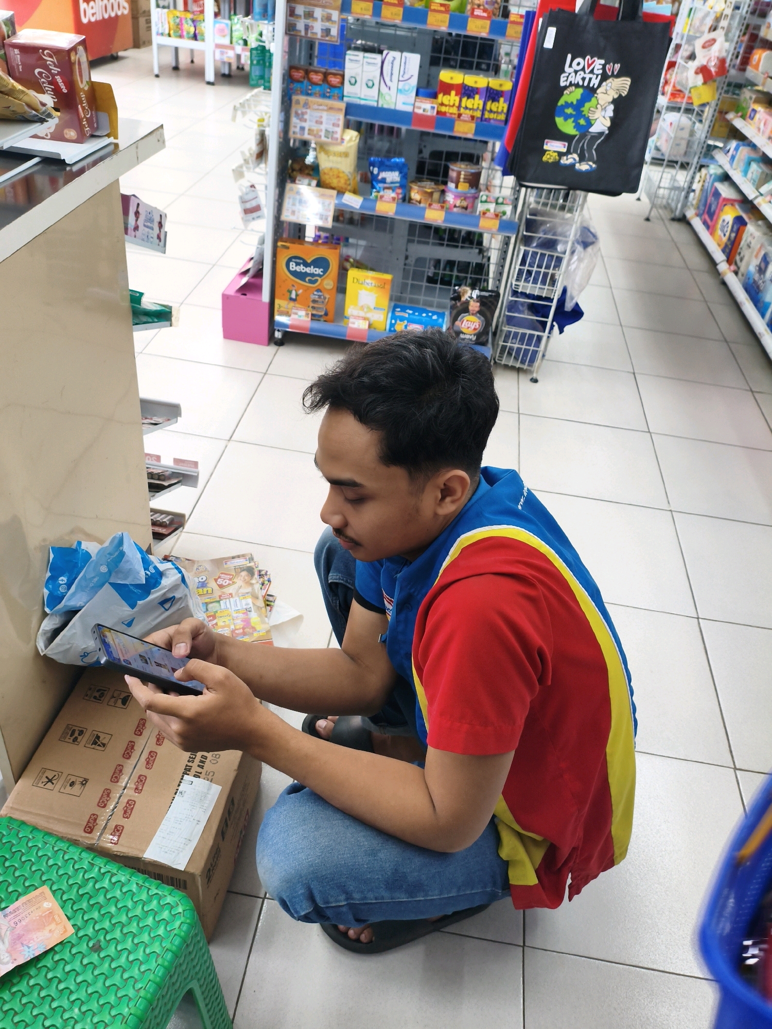 A young man is kneeling on the tiled floor of a convenience store in Sumberejo Kulon, Indonesia, looking at his smartphone. He is wearing a red and yellow polo shirt and blue jeans. A cardboard box is on his right, and a blue plastic bag is on his left. Shelves stocked with various products are visible in the background, including bags of chips and a tote bag with "LOVE EARTH" printed on it. A green plastic stool with a banknote on it is in the foreground.