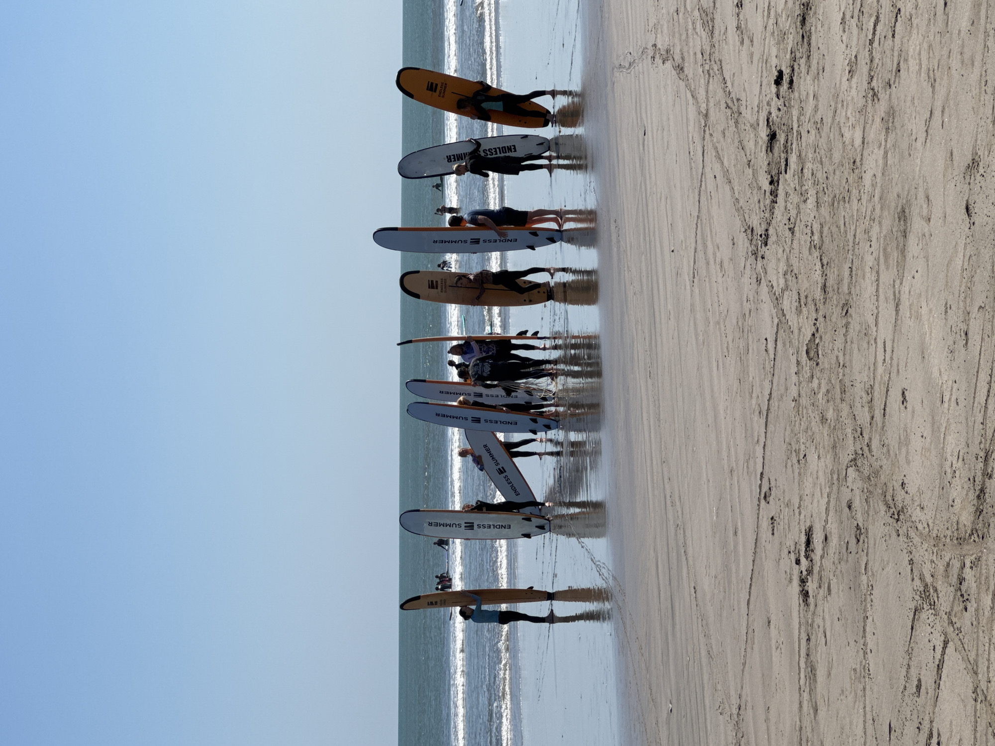 A group of approximately ten individuals, some of whom are wearing wetsuits, stand on a wet, sandy beach, holding a variety of surfboards upright. The surfboards are positioned in a rough, semi-circular formation. The individuals are facing the ocean, which has moderate waves breaking near the shore. The sky is clear and blue. The location is the beach of Kuta, Indonesia.