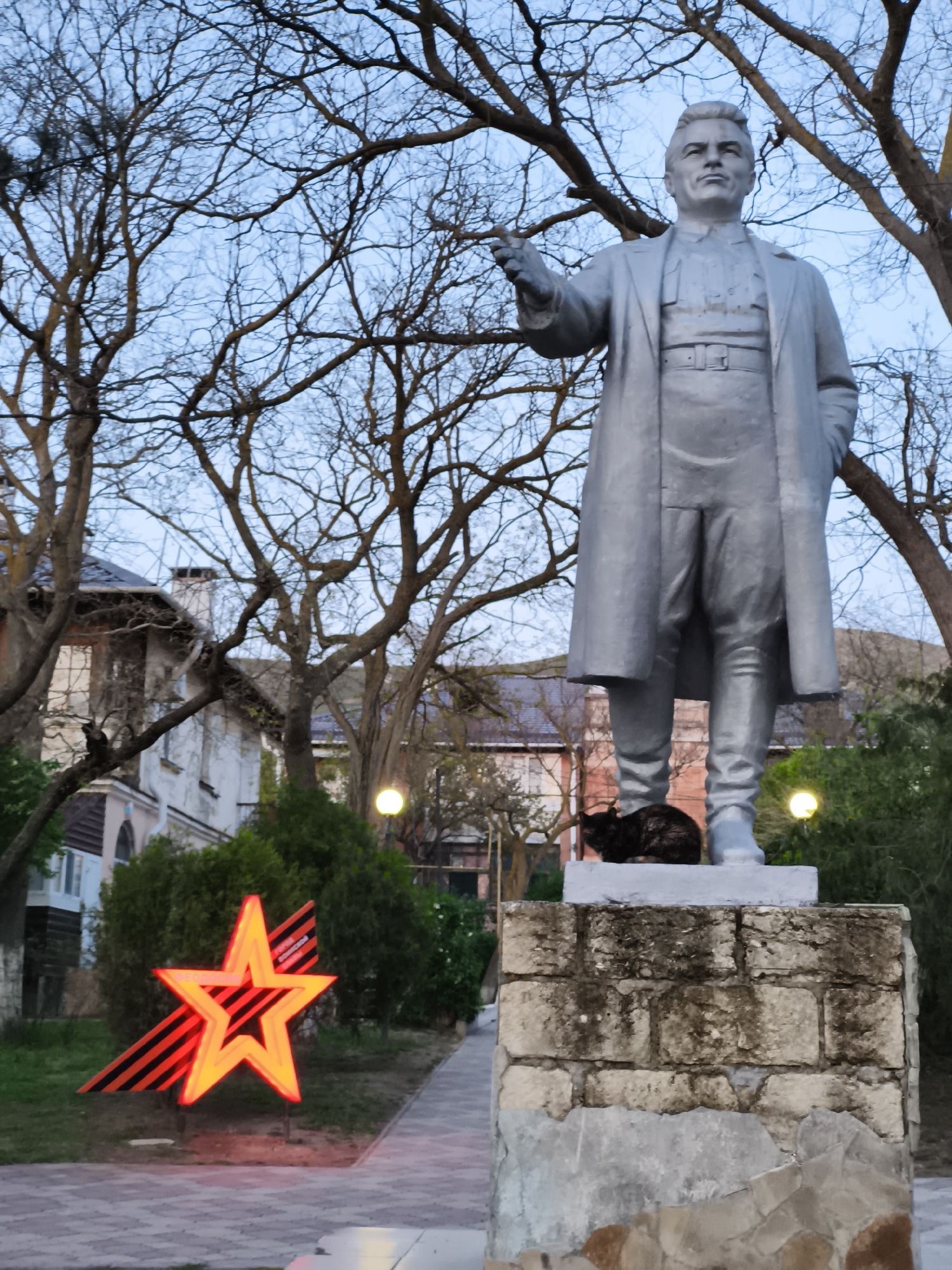 A statue of a man in a long coat stands in a park in Орджоникидзевский поселковый совет, Ukraine. The statue is made of stone or metal and stands on a brick pedestal. A dark-colored cat is sitting on the pedestal at the base of the statue. In the background, there is a building with several stories and bare trees. To the left of the statue, there is a red star shape with black and orange stripes behind it.