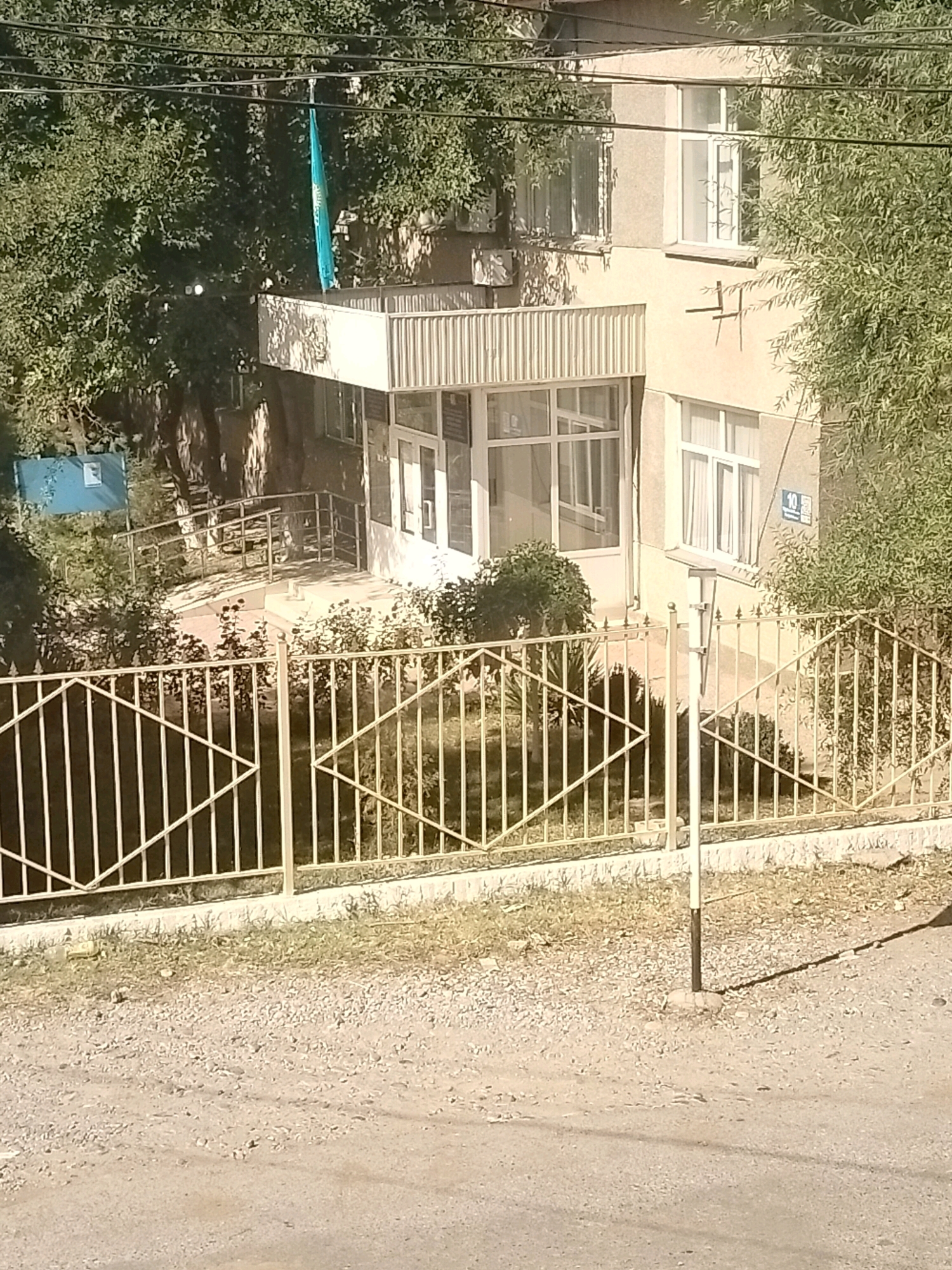 The image captures a street view in the city of Kölkent, Kazakhstan. In the foreground, a gravel road transitions to a dirt path leading to a light-colored metal fence with a diamond pattern. Beyond the fence, a small patch of dry grass and a few bushes are visible. The main subject is a multi-story building with a light beige facade. The building has several windows, some with curtains, and a covered entrance area with a glass enclosure and a ramp with metal railings leading to it. A blue sign with some text is partially visible near the ramp. A flag, appearing to be light blue with some emblem, is hanging from a wire strung between trees and the building. Several power lines are also visible in the upper part of the image. The environment suggests a sunny day with shadows cast by the trees and the building.